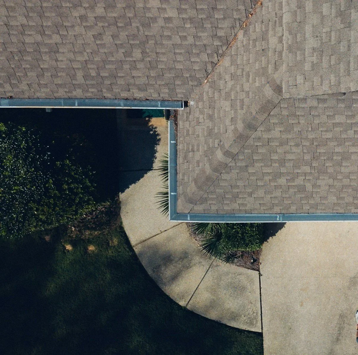 An aerial view of a house's roof with shingles, showing the gutter along the edge of the roof and part of the yard with a curved sidewalk and some bushes.