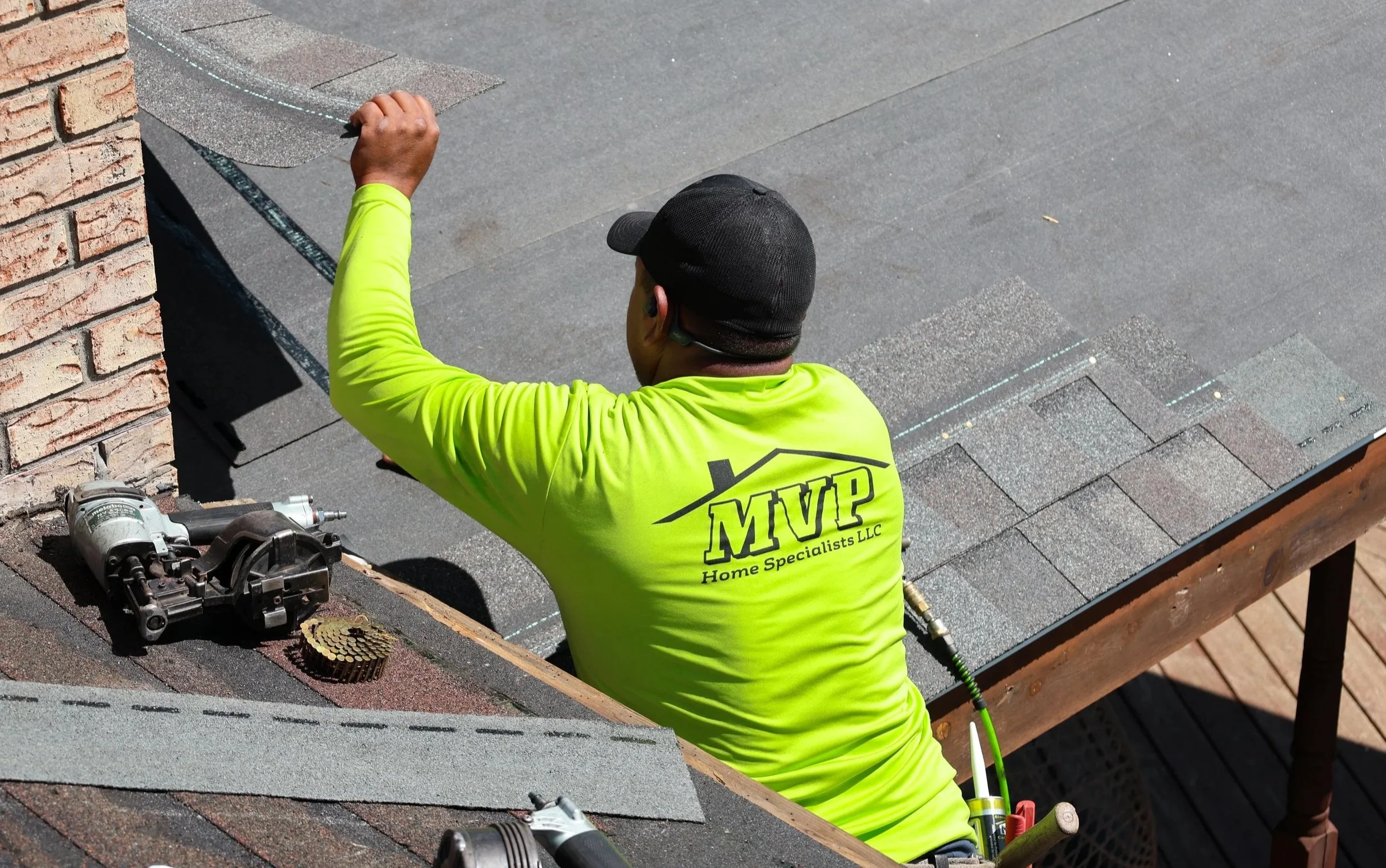 A worker installing roofing shingles on a house. The worker is wearing a neon yellow shirt with the logo 'MVP Roof Pros' and a black baseball cap, and is using a tool to work on the roof.