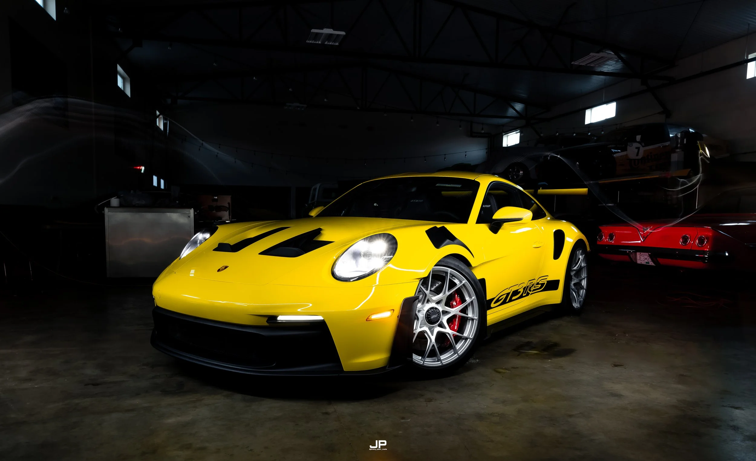 Yellow Porsche 911 GT3 RS sports car parked indoors under dim lighting, with black decals and red brake calipers visible through the wheels, other vehicles in the background.