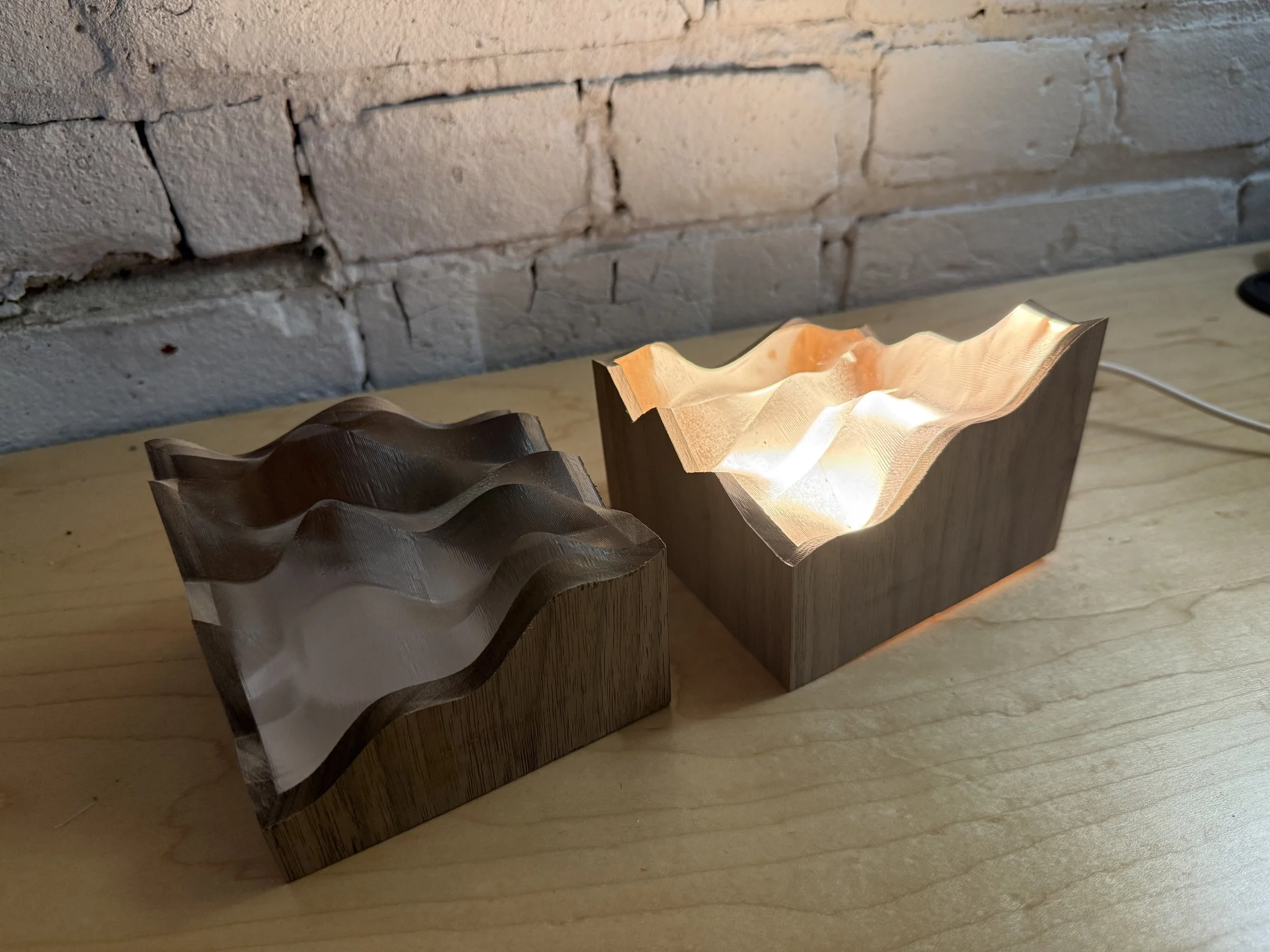Two wooden lamps with wavy, carved tops, turned on and illuminating a wooden surface, against a white brick wall background.