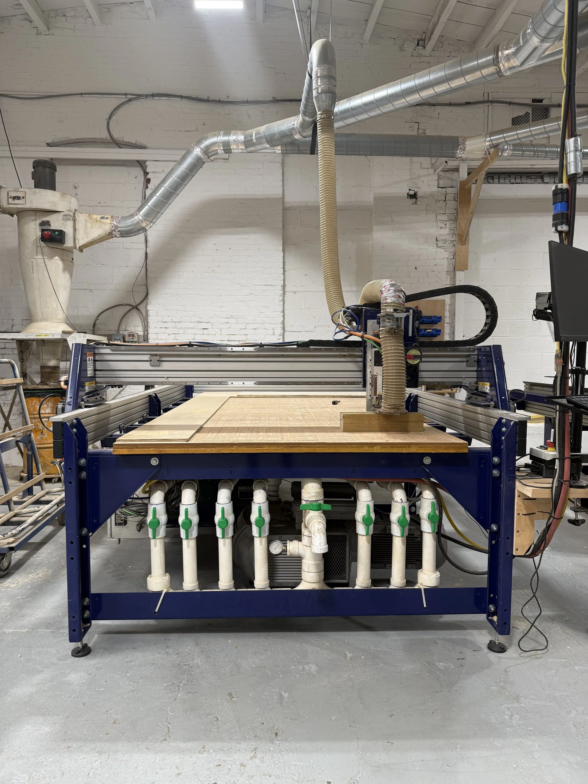 A CNC wood carving machine in a workshop, with a computer monitor and various pipes and ventilation systems in the background.