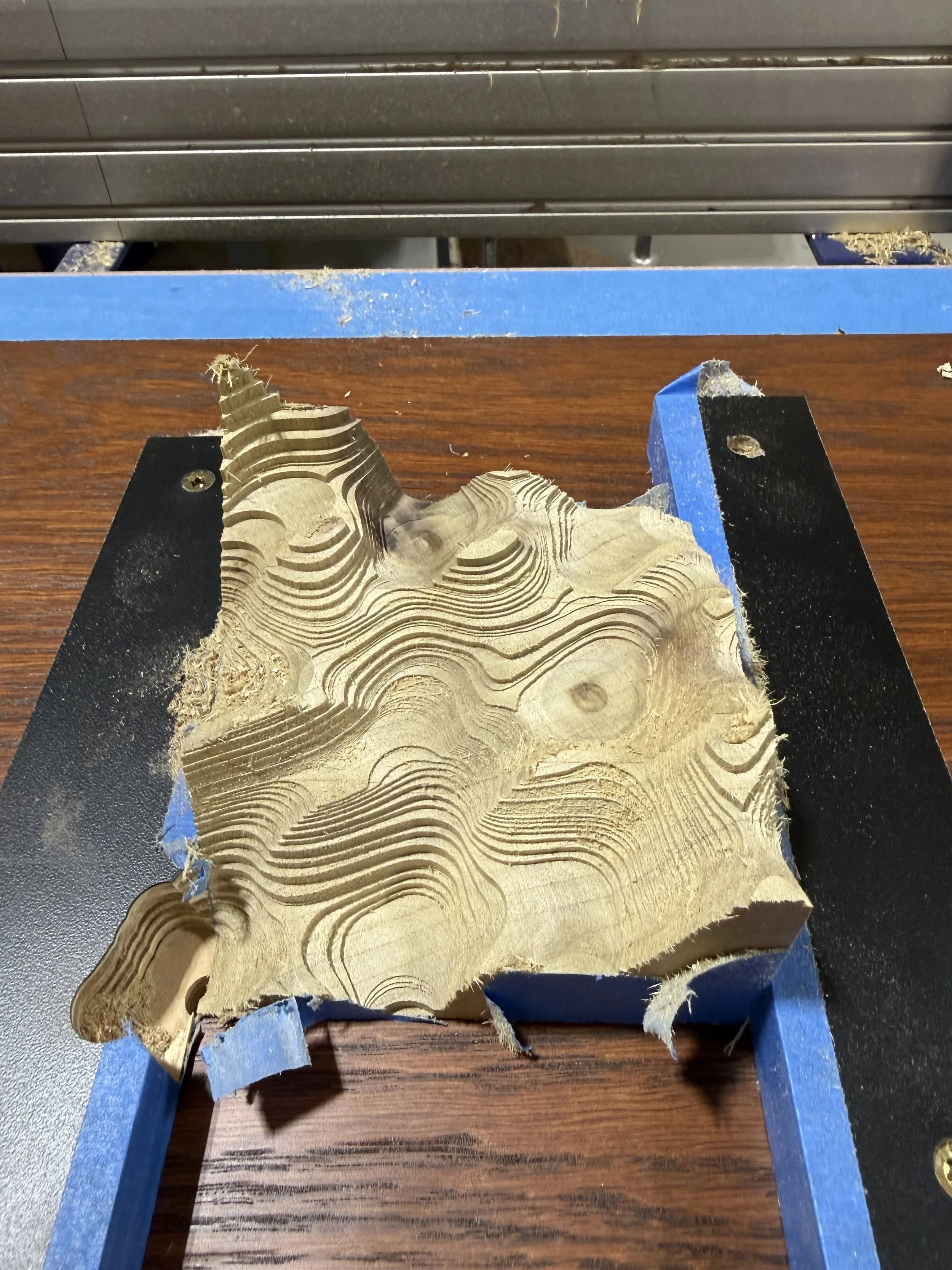 Close-up of a piece of layered wood, possibly wood veneer, clamped to a work surface with blue painter's tape on the edges.