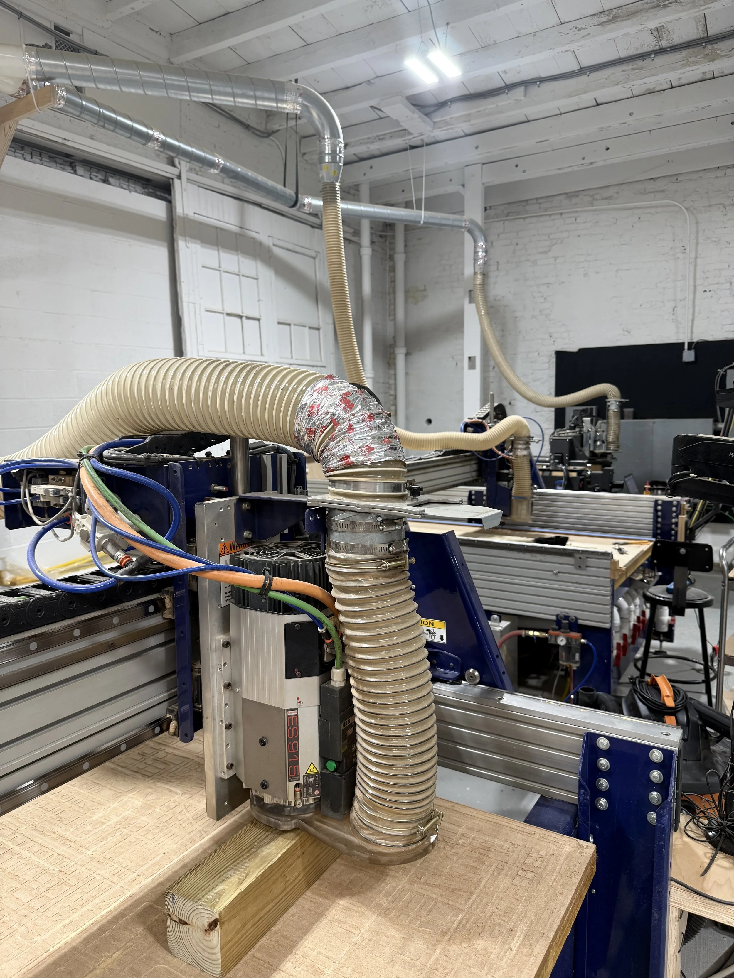 Industrial laser machine with ventilation hoses in a workshop with white brick walls and ceiling.