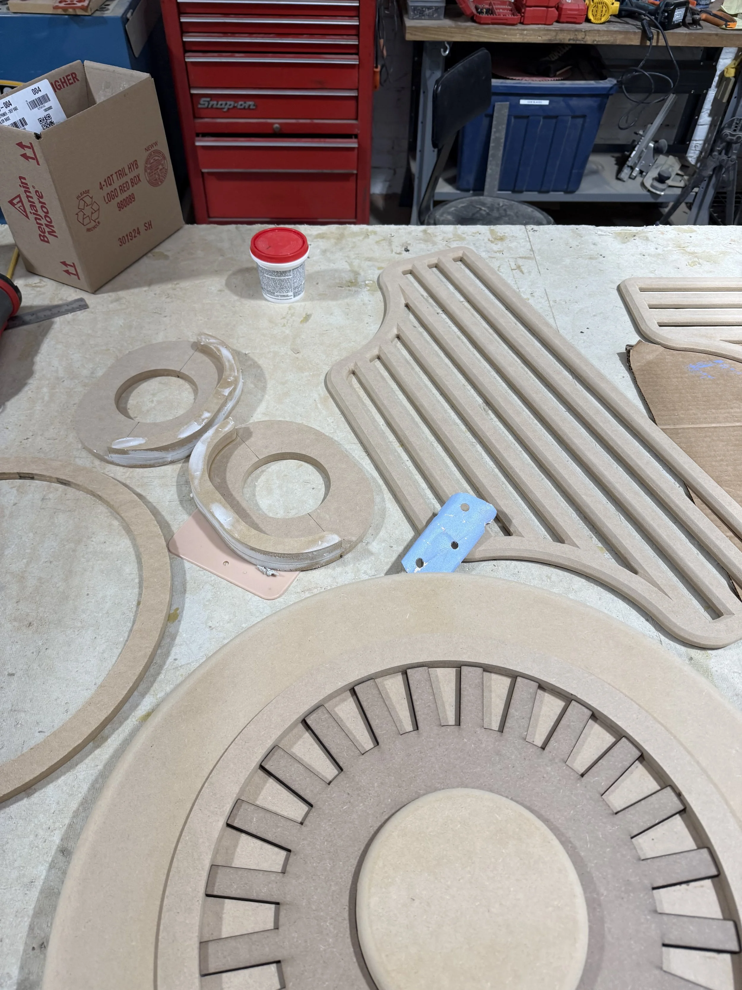 Wooden clock or gear parts on a workshop workbench with various tools and storage drawers in the background.