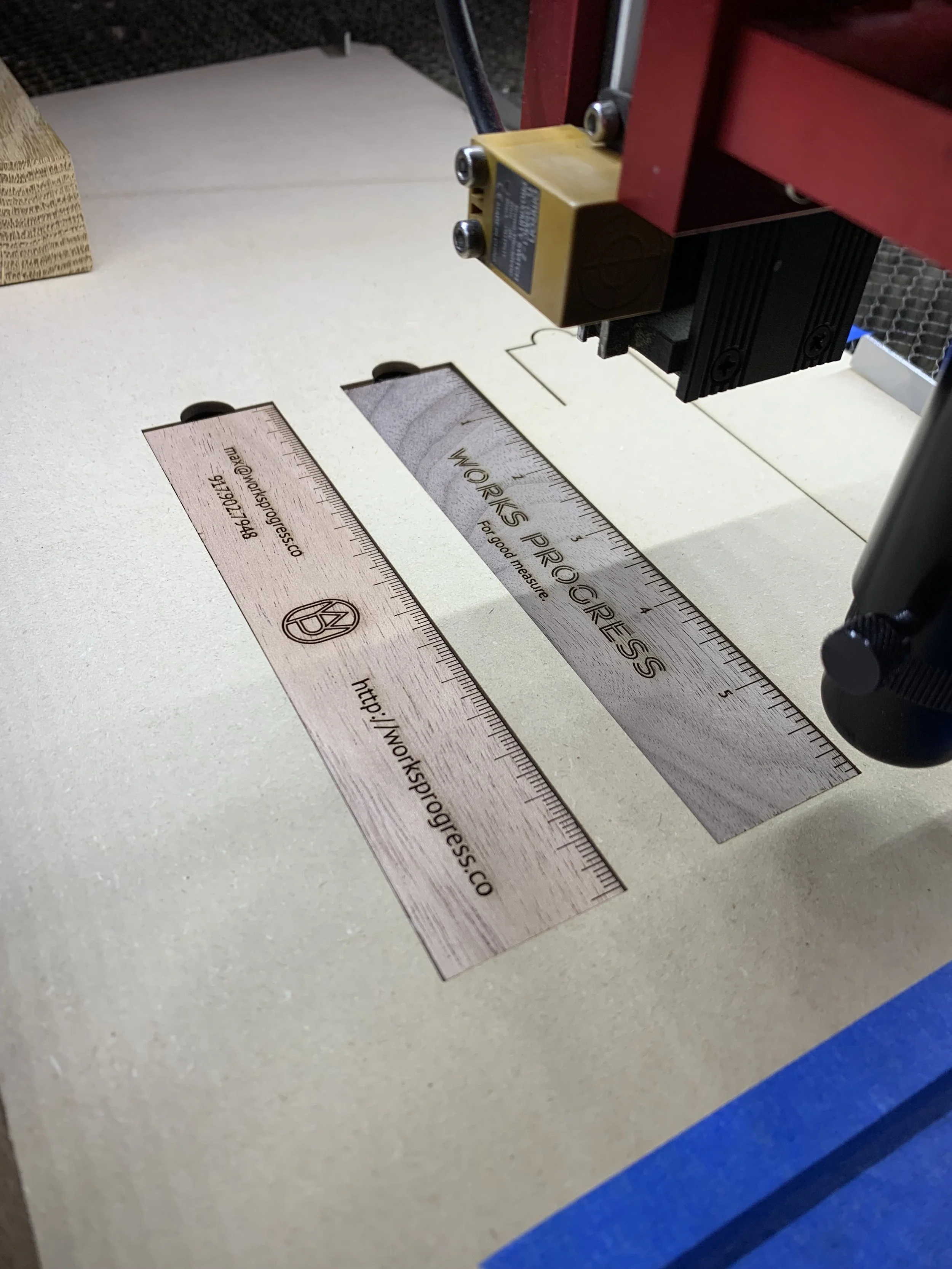 A laser engraving machine carving a wood-like design into a piece of material with rulers indicating measurements. The design includes the text "WORKS PROGRESS" and a URL, along with a VW logo.
