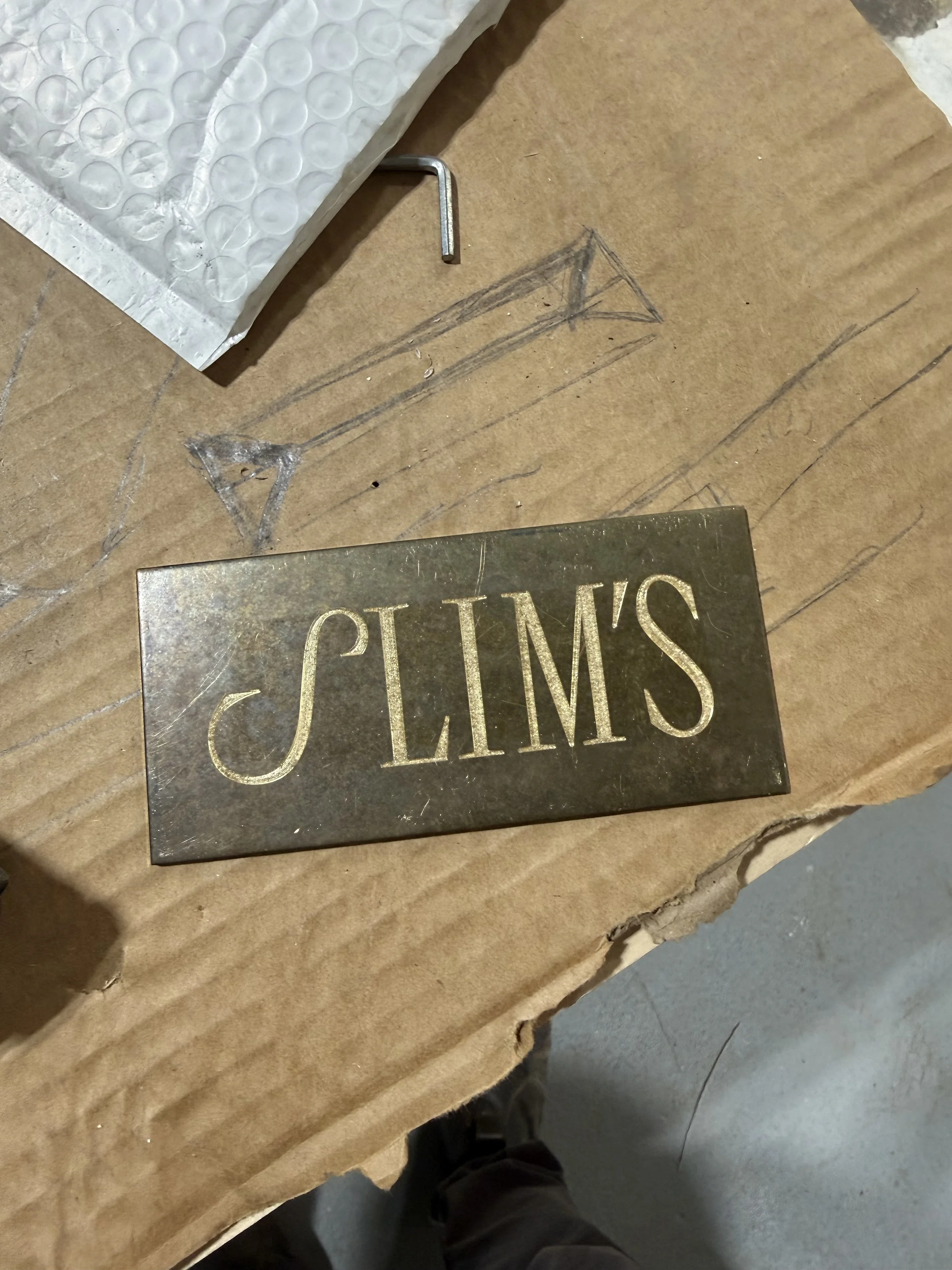 A metal plaque with the word 'SLIM'S' engraved on it lying on a workbench when viewed from above.