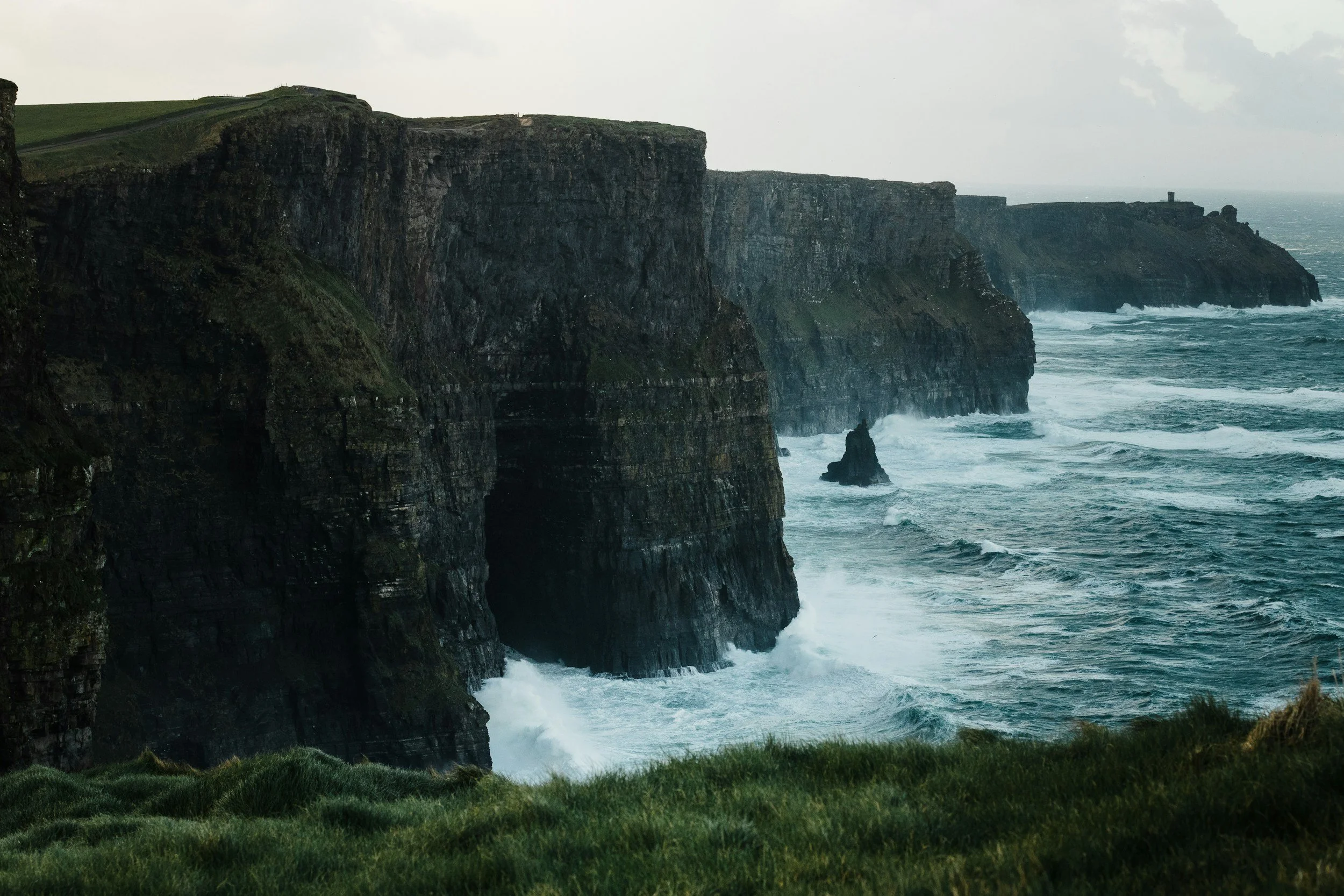 A tall cliffside, with crashing waves at the bottom of the cliff, a grey foggy sky behind the tall cliffs