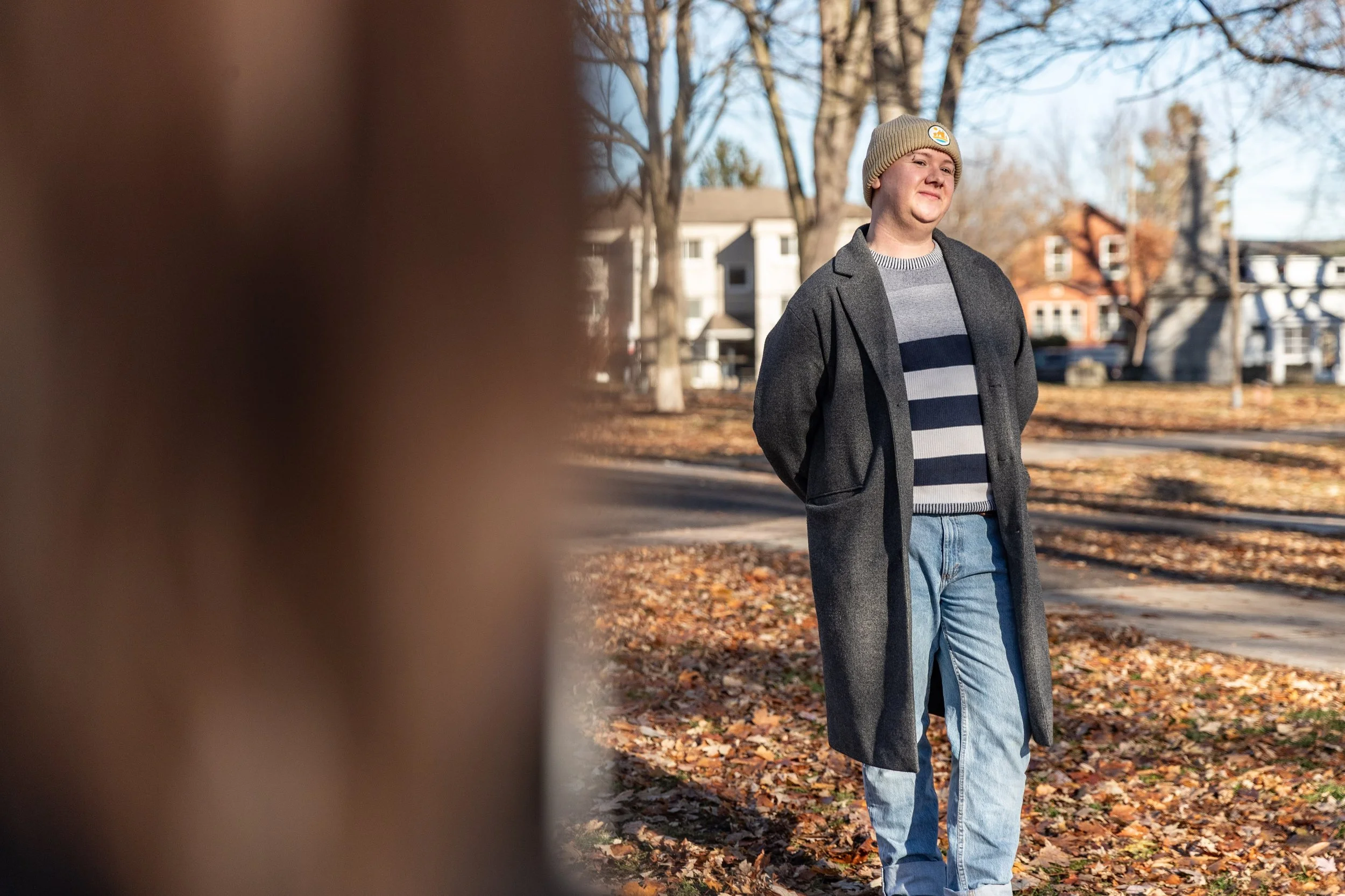 Man (pale complexion, grey wool, long coat, light-wash jeans, a white and blue striped sweater, wearing a beige tuque) smiling to someone off camera standing in a park, fall leaves covering the ground