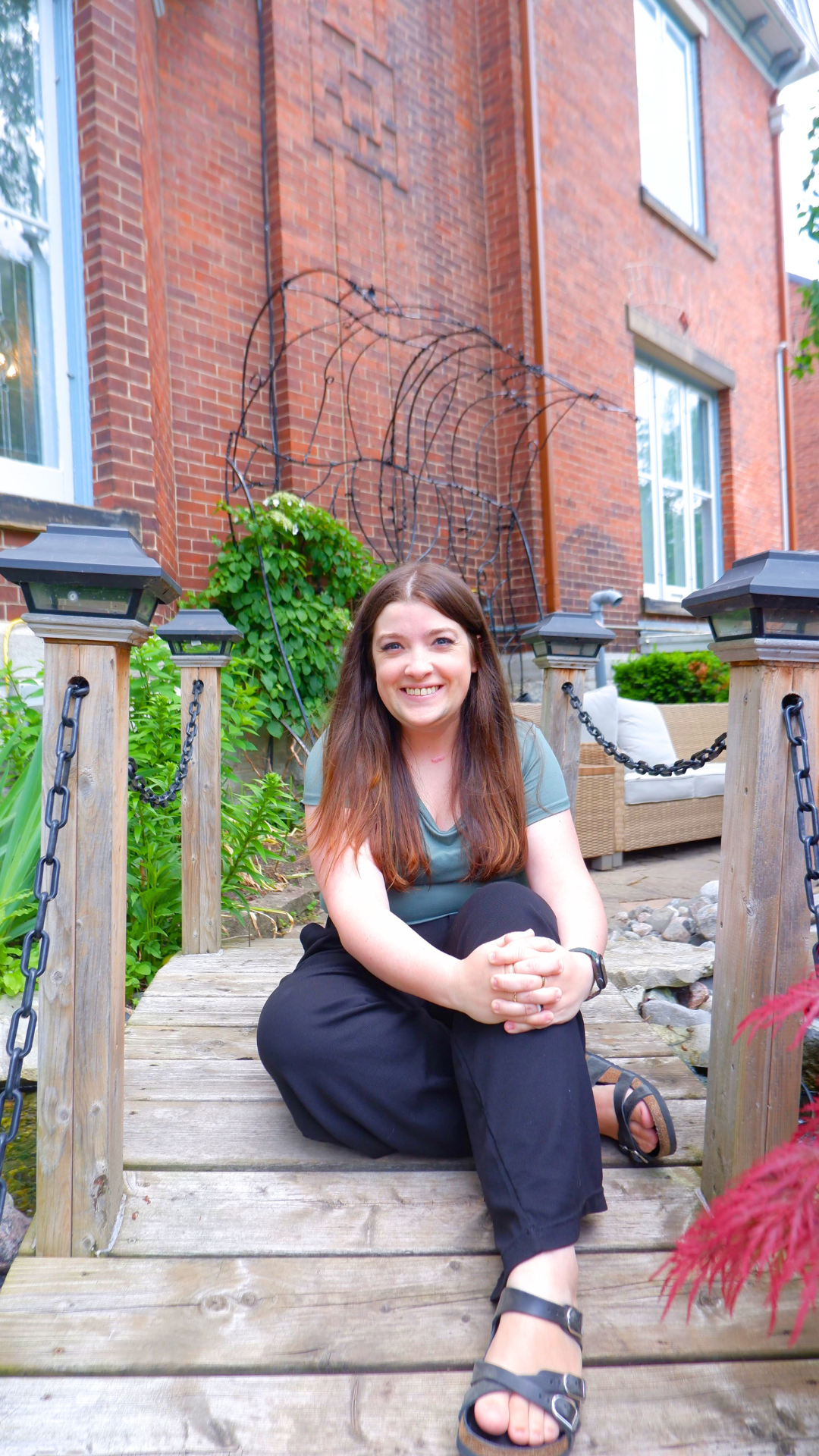 Erika (long, straight brown hair, wearing a muted sage green t-shirt, black work pants, black-strapped sandals) smiling at camera, sitting on a small wooden bridge in a zen garden, a red brick wall behind her with bright green foliage growing against