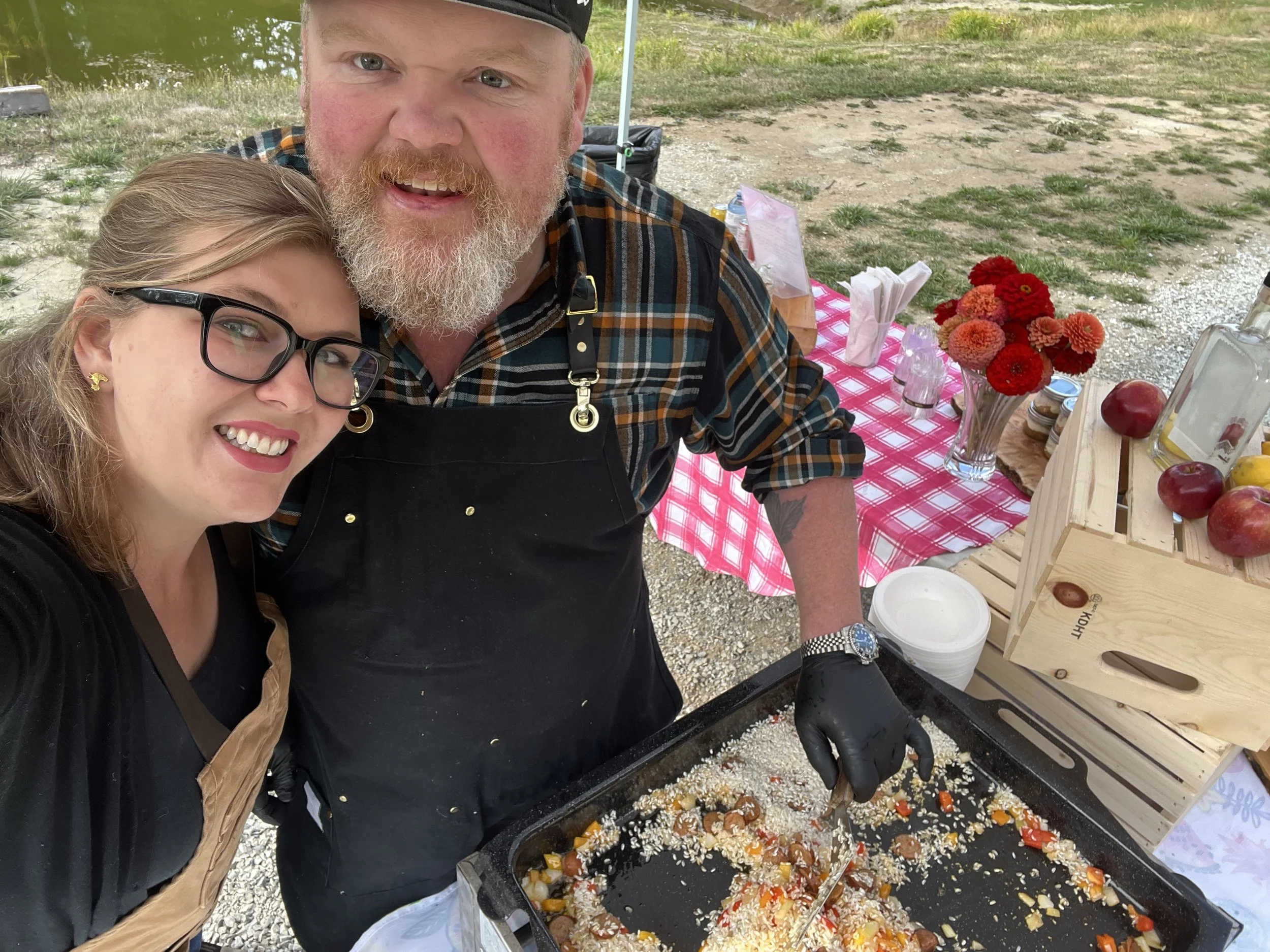 A woman and man smiling and posing for a selfie outdoors at a food stand. The man is cooking rice and vegetables on a griddle, wearing an apron and black glove. The stand has a red and white checkered cloth with flowers, apples, and condiments.