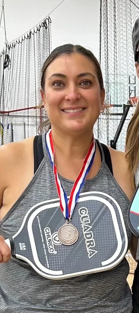 A woman smiling, wearing a medal around her neck, holding a large bar of soap with the words Quadar and Churco, at an indoor sports event.