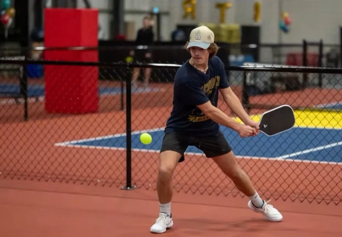 A person playing pickleball in an indoor sports facility, preparing to hit a yellow ball with a paddle, wearing a baseball cap, navy T-shirt, shorts, and white sneakers.