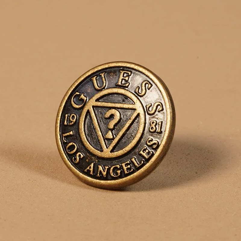 A circular pin with the words 'GUESS LOS ANGELES 1981' surrounding a stylized triangle and question mark in the center.