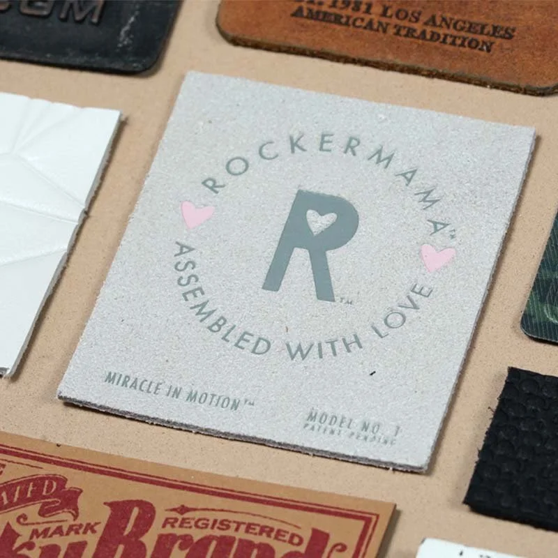 Close-up of a square bathroom tile with the words "ROCKER MAMA" and "ASSEMBLED WITH LOVE" around a large letter R with a small heart in the middle, and two small pink hearts on either side.