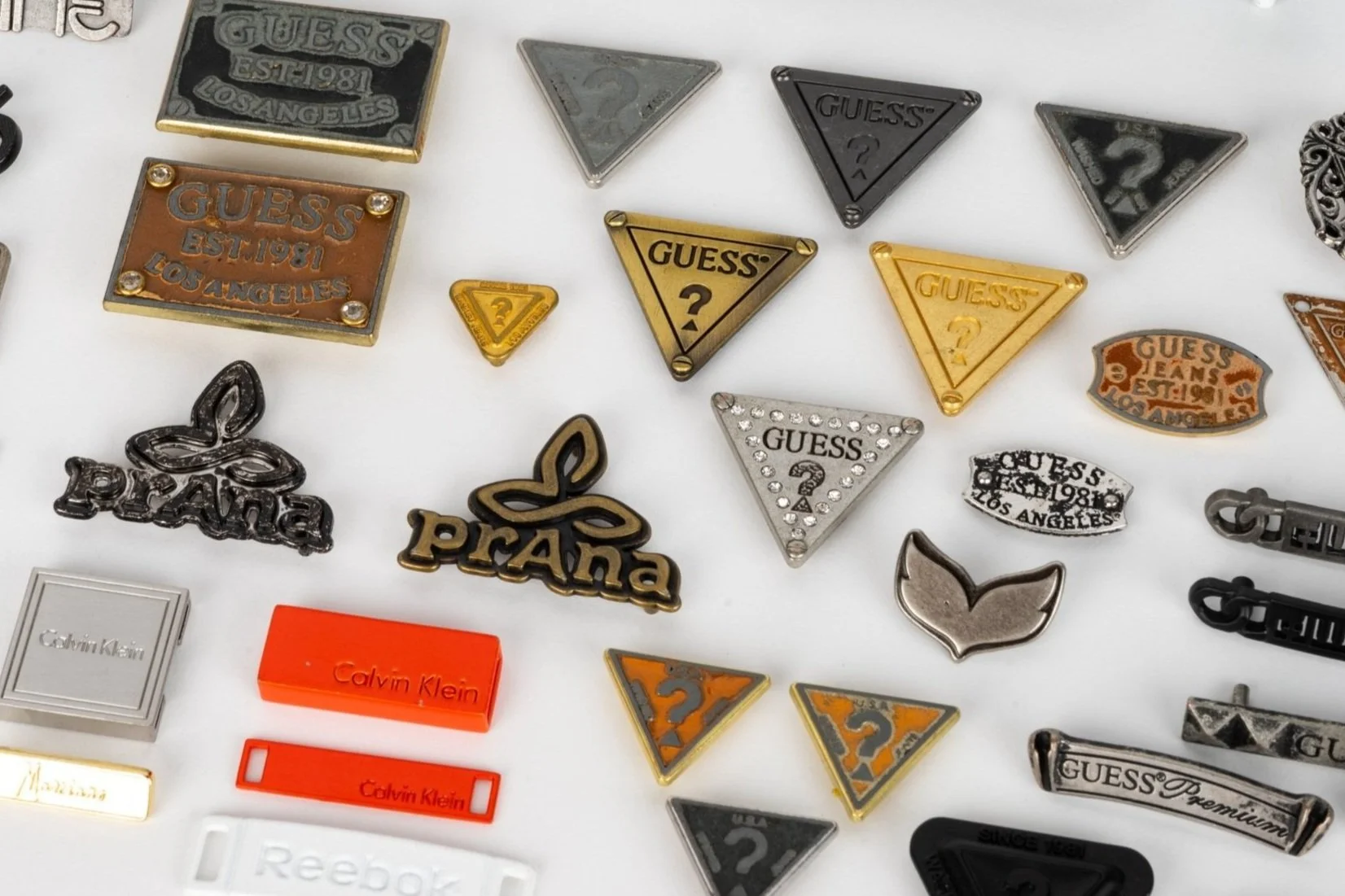 A collection of various designer pins and patches on a white surface, including Guess, Prada, Calvin Klein, and other brand logos and symbols.