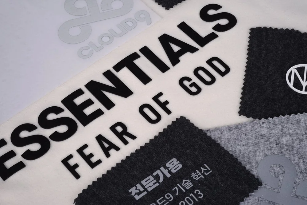 A collection of fabric swatches and stickers, with the prominent text "ESSENTIALS FEAR OF GOD" in black on a white background.