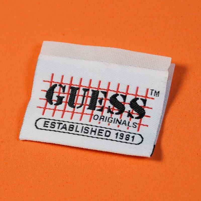 Close-up of a white fabric label with the Guess logo, black text, red grid design, and orange background.