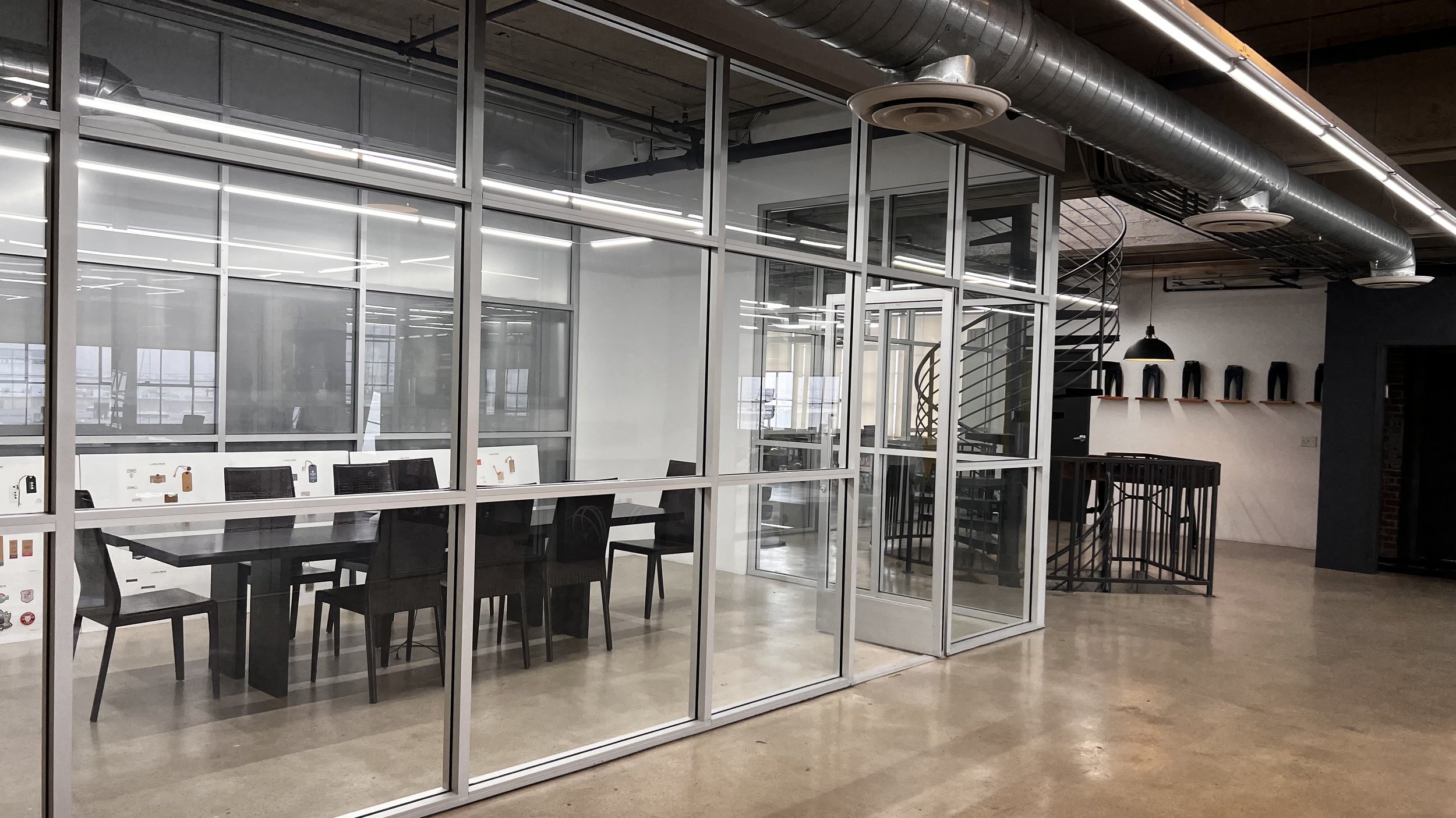 Modern office conference room with glass walls, black chairs, a long table, and a spiral staircase in the background.