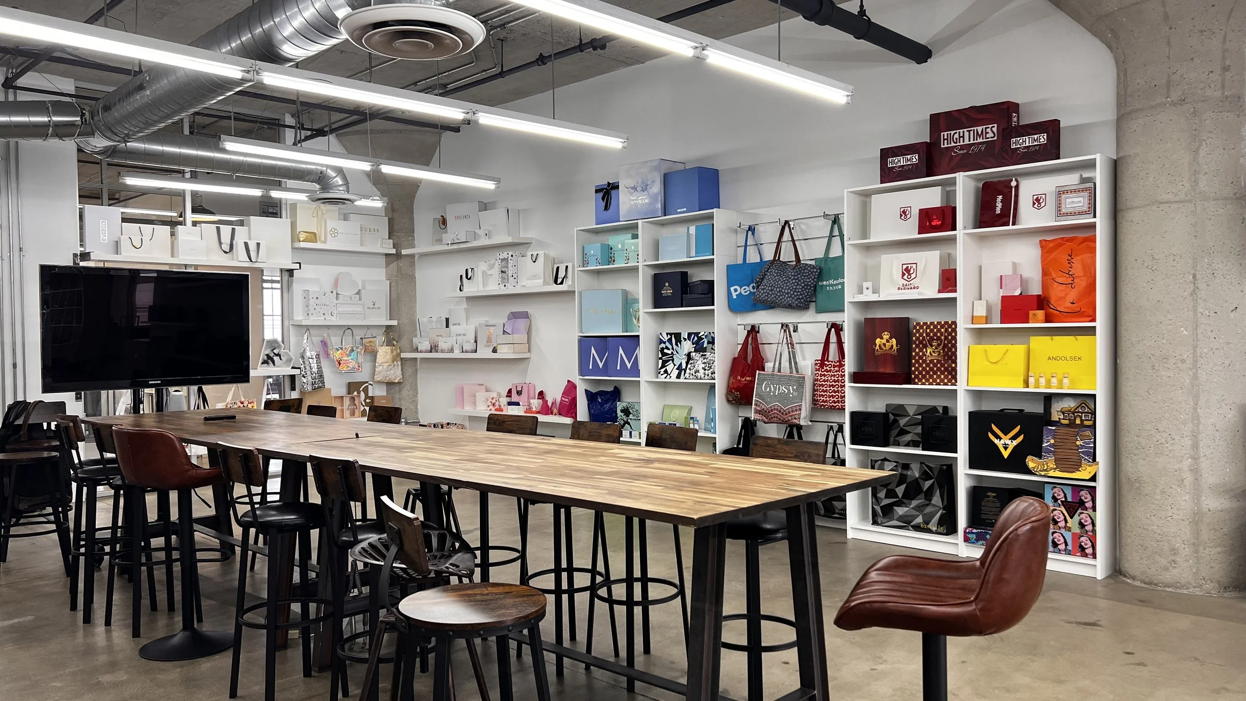 An interior view of a modern showroom with a large wooden table, surrounded by various chairs and stools, and shelves filled with colorful bags, boxes, and souvenirs, and a large TV screen on the left.