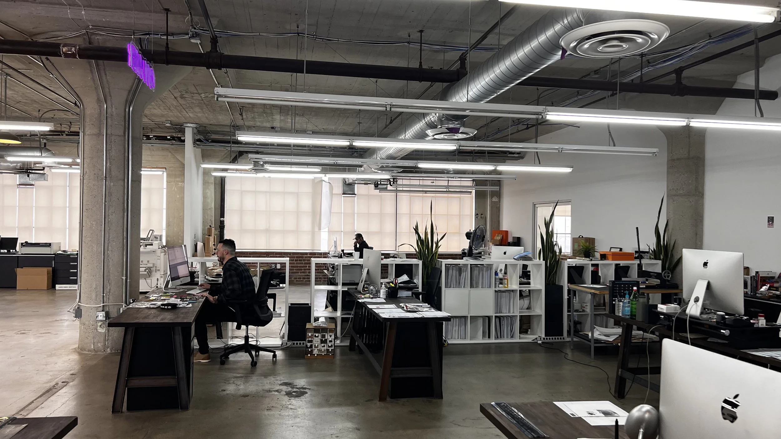 Open office area with multiple workstations, desktop computers, and employees working. Industrial-style space with exposed concrete ceiling, air ducts, and large windows with blinds.