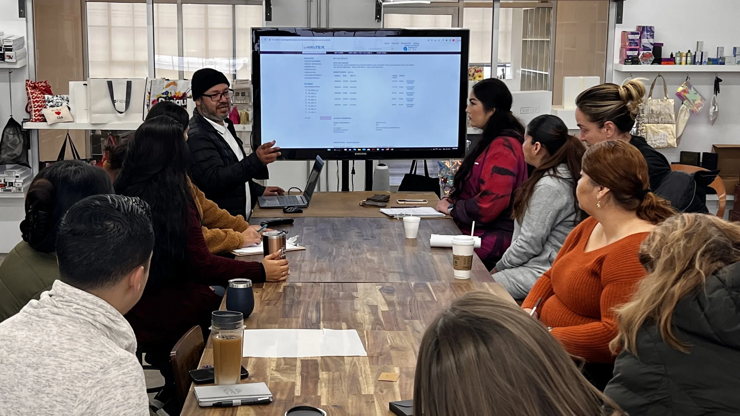 A group of people gathered around a wooden conference table, listening to a presenter who is pointing at a large screen displaying a spreadsheet or data table during a meeting or workshop.