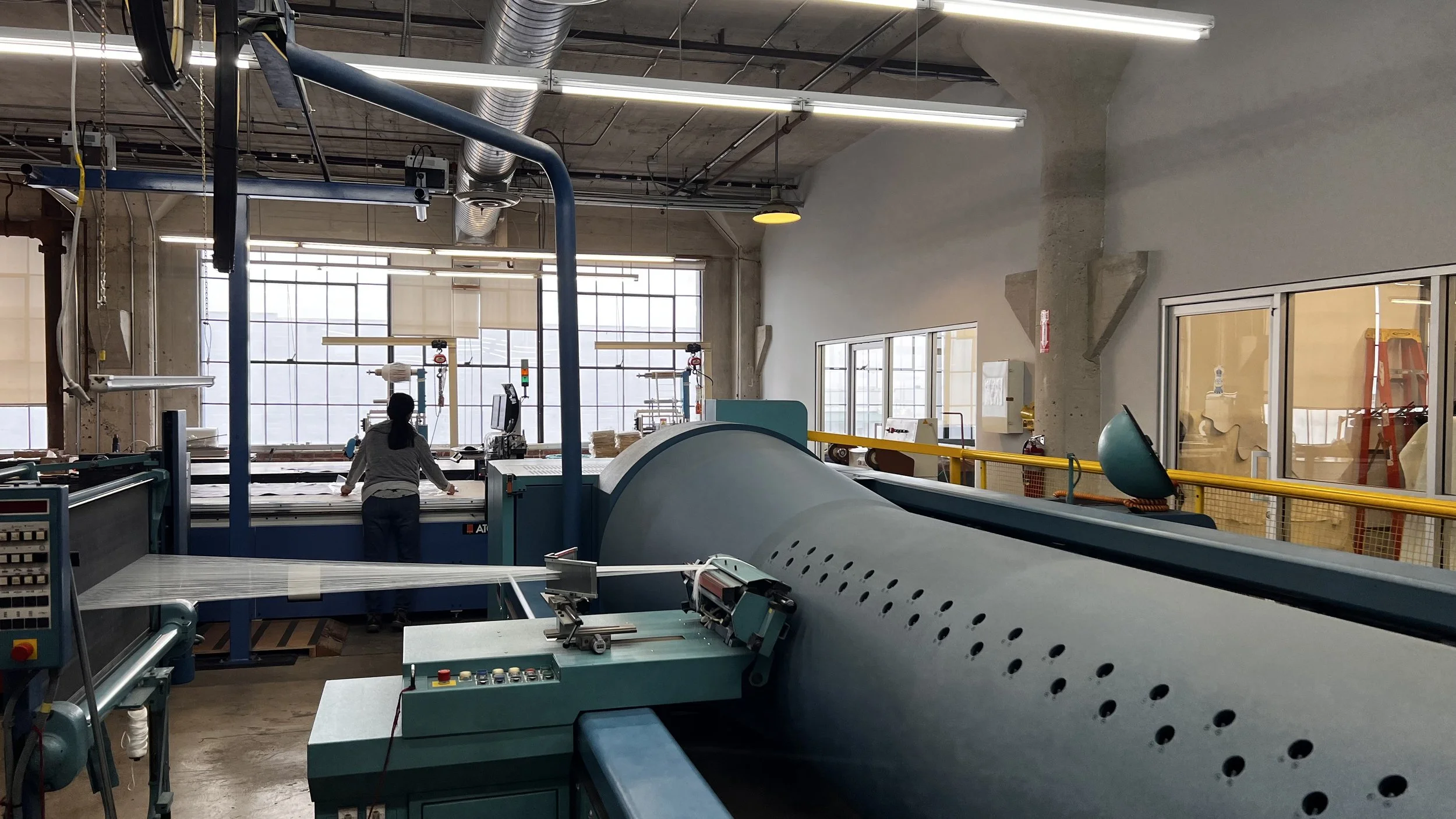 A person operating machinery in an industrial workshop with large windows, metal pipes, and various equipment.