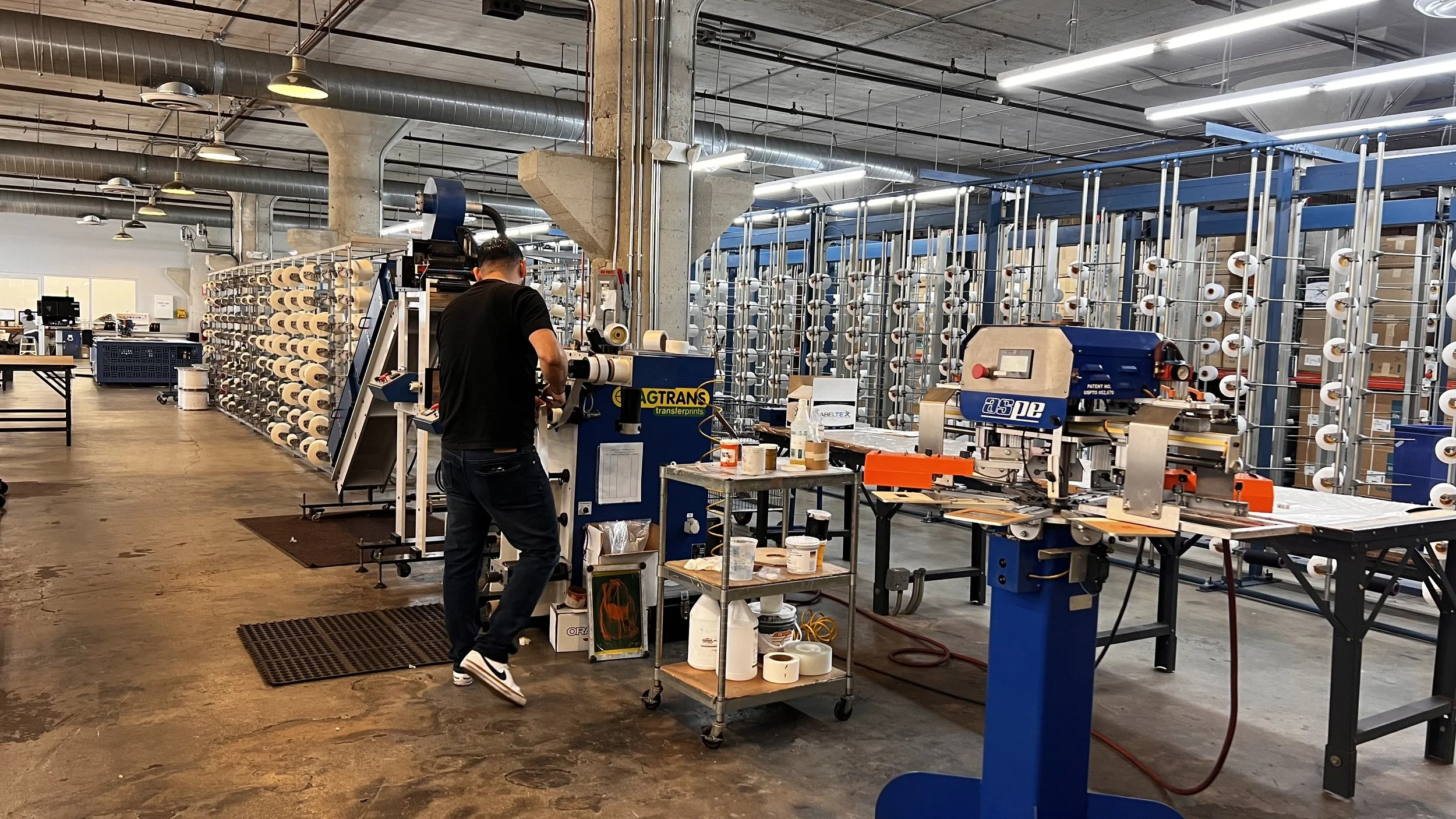 Worker operating machinery in a textile manufacturing factory with numerous spools of material on racks in the background.