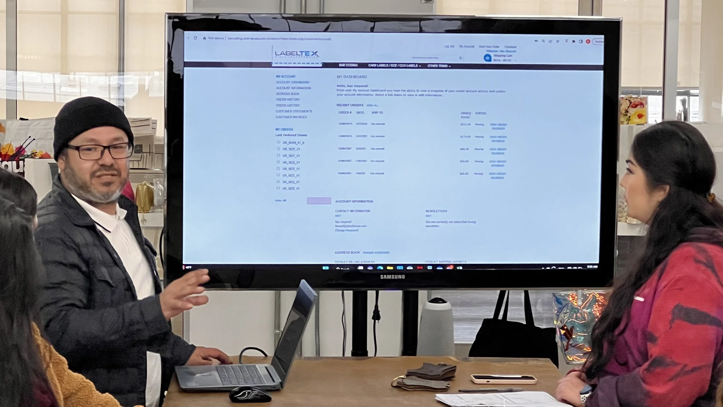 People attending a presentation or training session in a public space, with a man standing and gesturing, and a woman listening, next to a laptop and a large screen displaying a website interface.