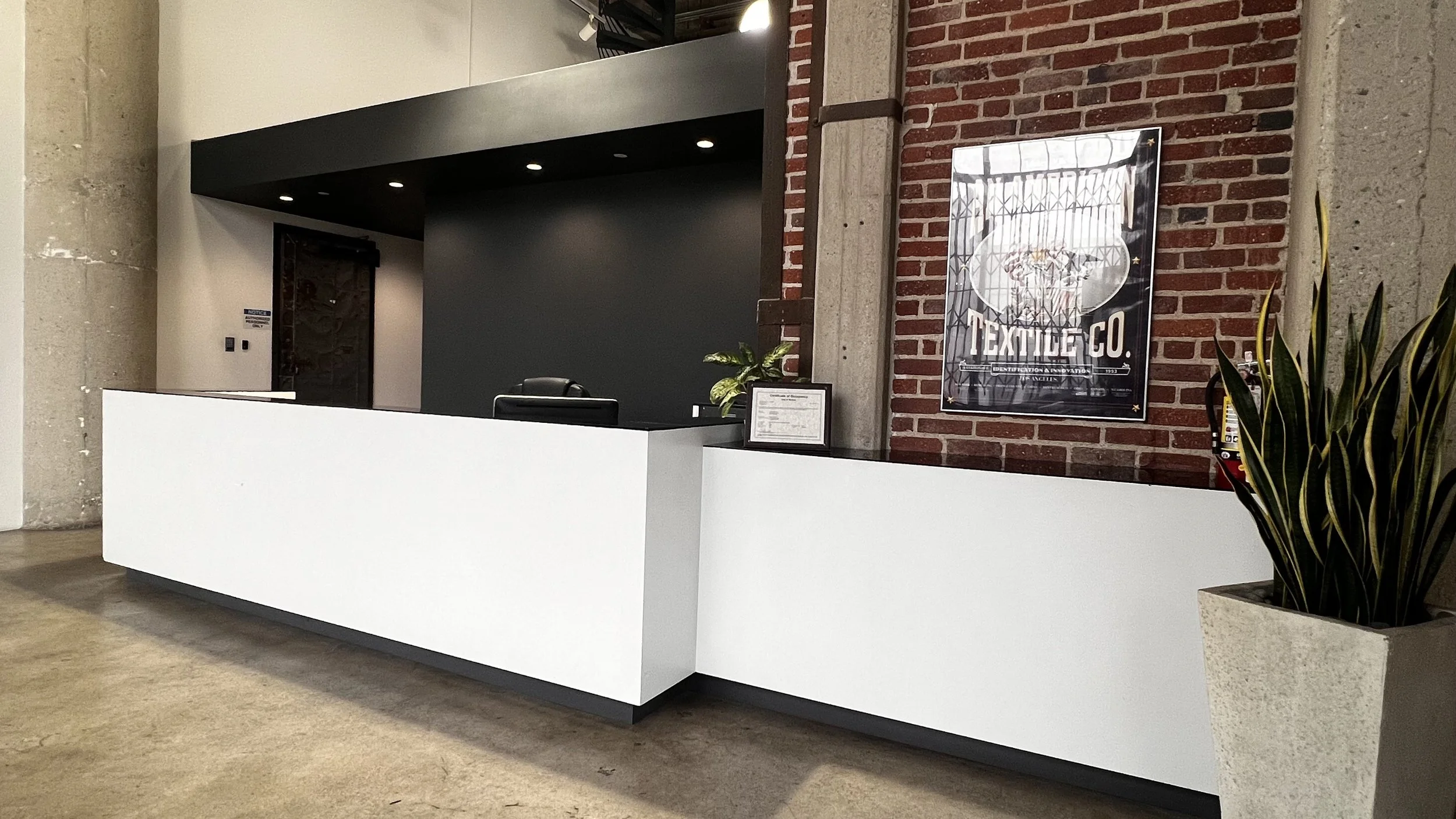 Modern reception desk with a black overhead canopy, a dark accent wall behind, exposed brick wall on the side, a framed logo or sign, and a potted plant in the foreground.