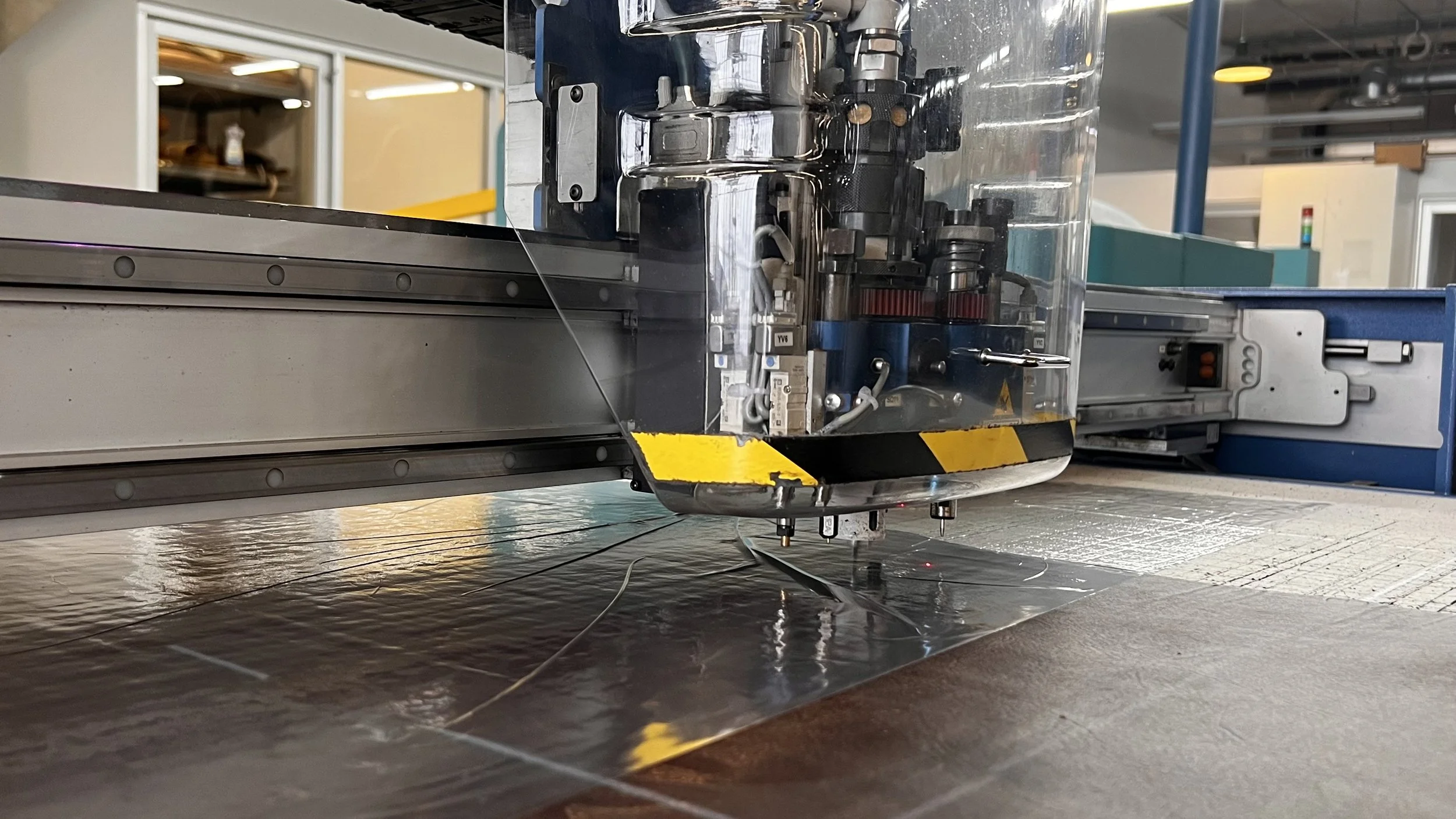 Close-up of a scientific or industrial machine with metal components, wires, and a transparent protective cover, working on a reflective surface in a modern laboratory or manufacturing environment.