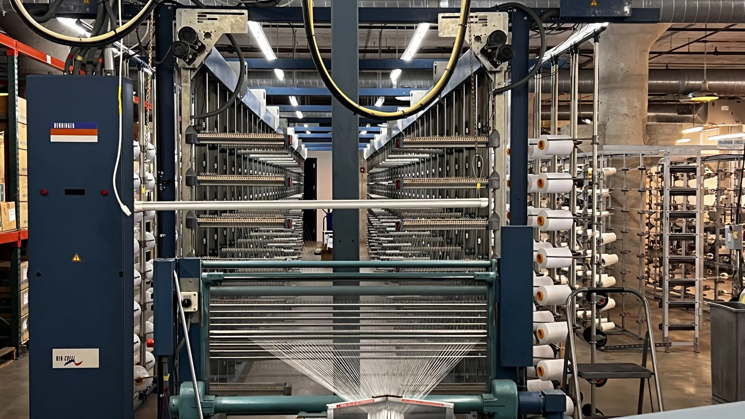 Industrial textile knitting machine in a factory, with multiple spools of white yarn and various mechanical parts.