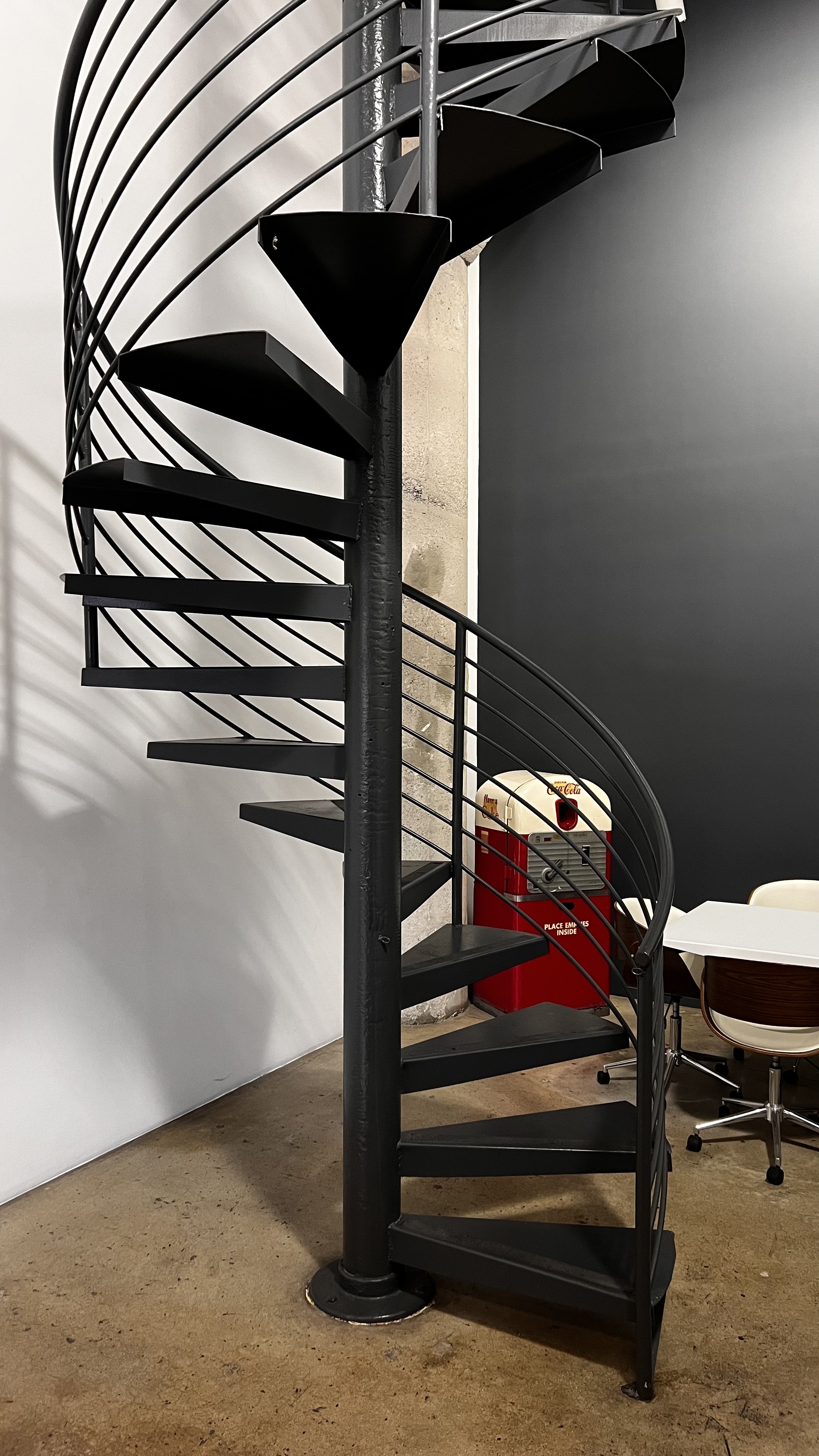 Black metal spiral staircase with a railing, located next to a white and black wall, with a red fire hydrant box and a table with white and wooden chairs nearby.