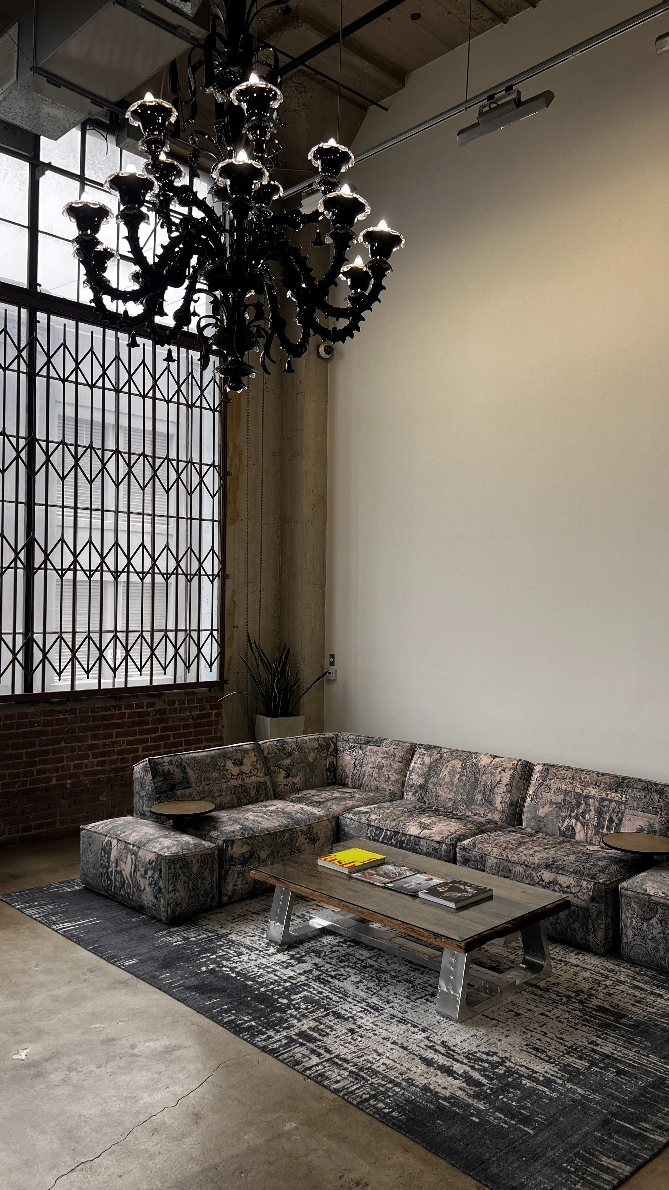 A modern industrial-style lounge area with a large floral-patterned couch, a rustic wooden coffee table with magazines, a black chandelier, large grid-style windows, a potted plant, and a textured rug on a concrete floor.