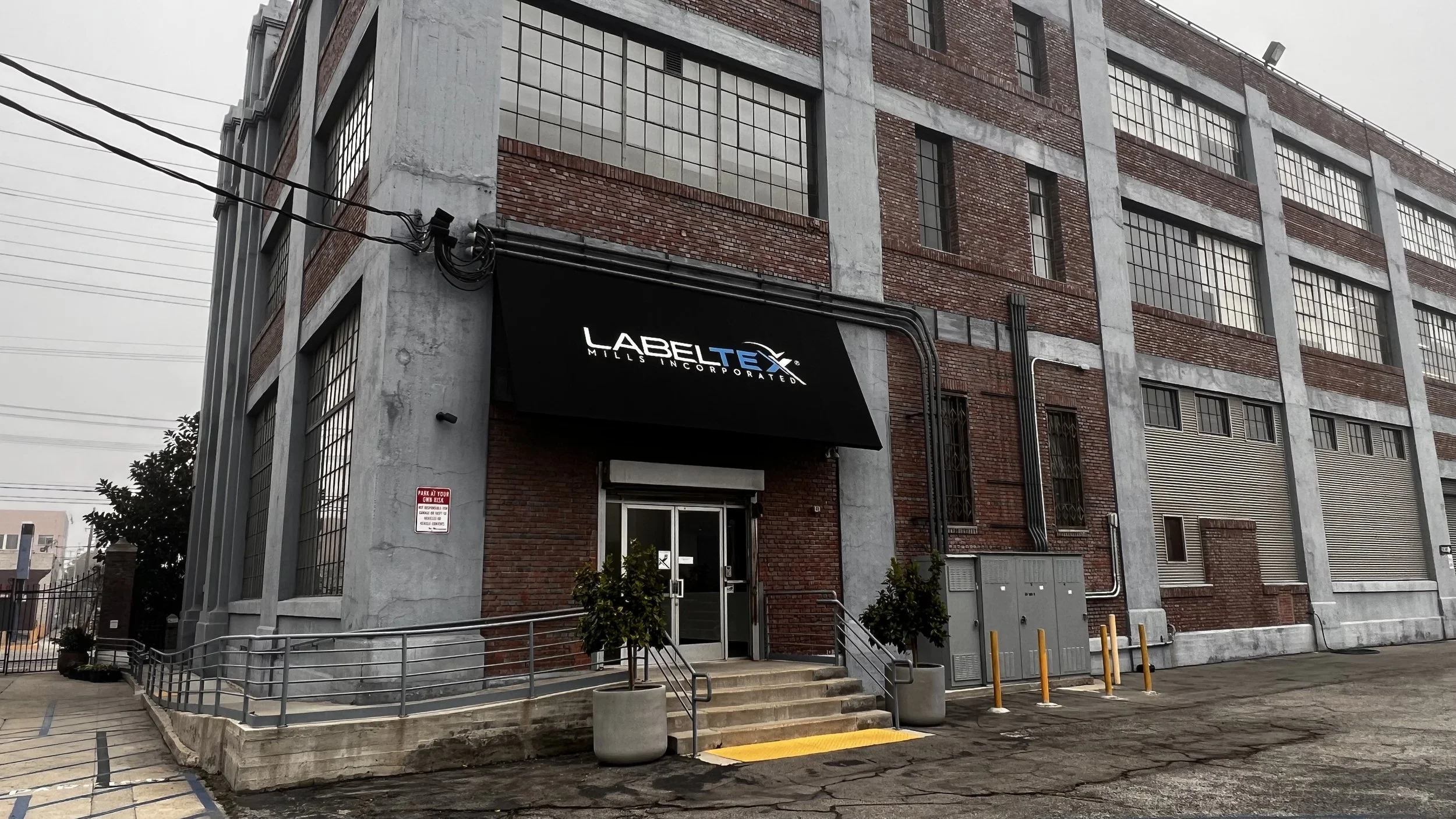 Exterior of an industrial-style brick building with a black awning that reads 'LABEL TEX MILL INCORPORATED.' The entrance has steps, a wheelchair ramp, and potted plants on either side.