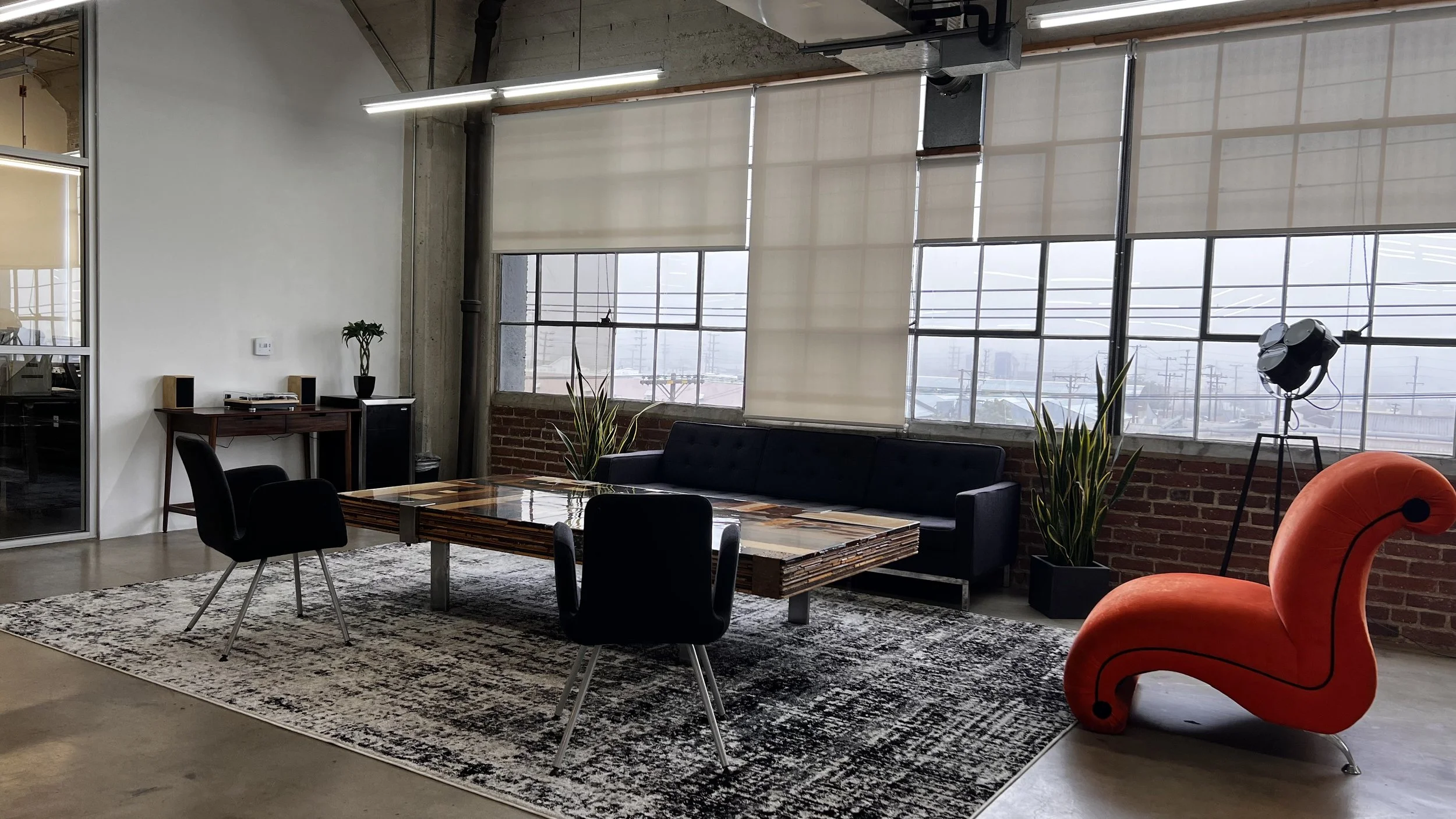 Modern office lounge area with a wooden coffee table, black sofa, black chairs, a red flamingo-shaped chair, tall floor lamp, potted plants, and large windows with blinds.