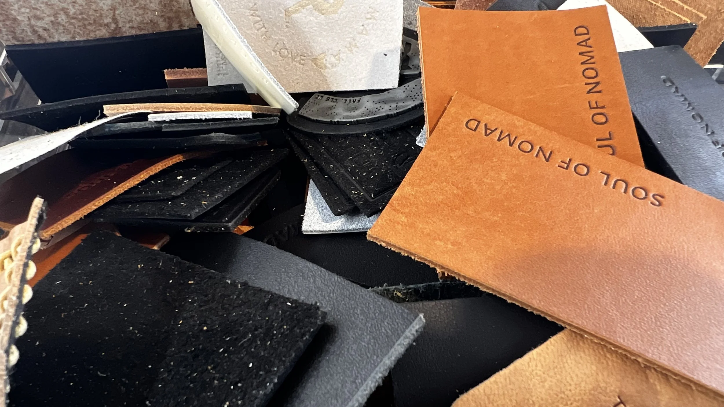 A pile of leather swatches in black, orange, tan, and gray with various textures and text, including 'SOUL OF NOMAD' on the orange swatches.