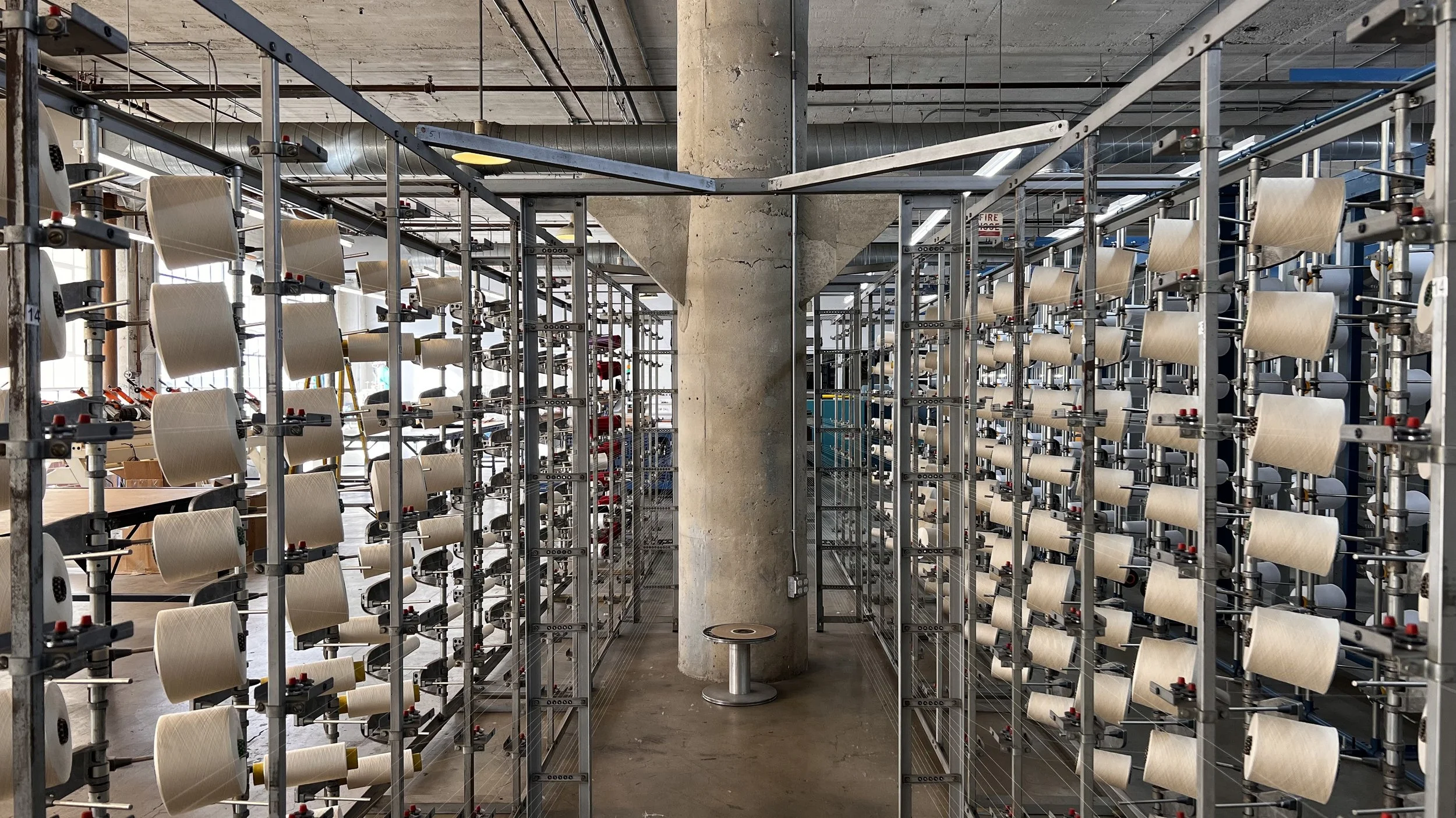 Industrial workspace with metal racks holding large spools of cream-colored thread, arranged in rows on either side of a concrete support pillar in a factory or workshop.