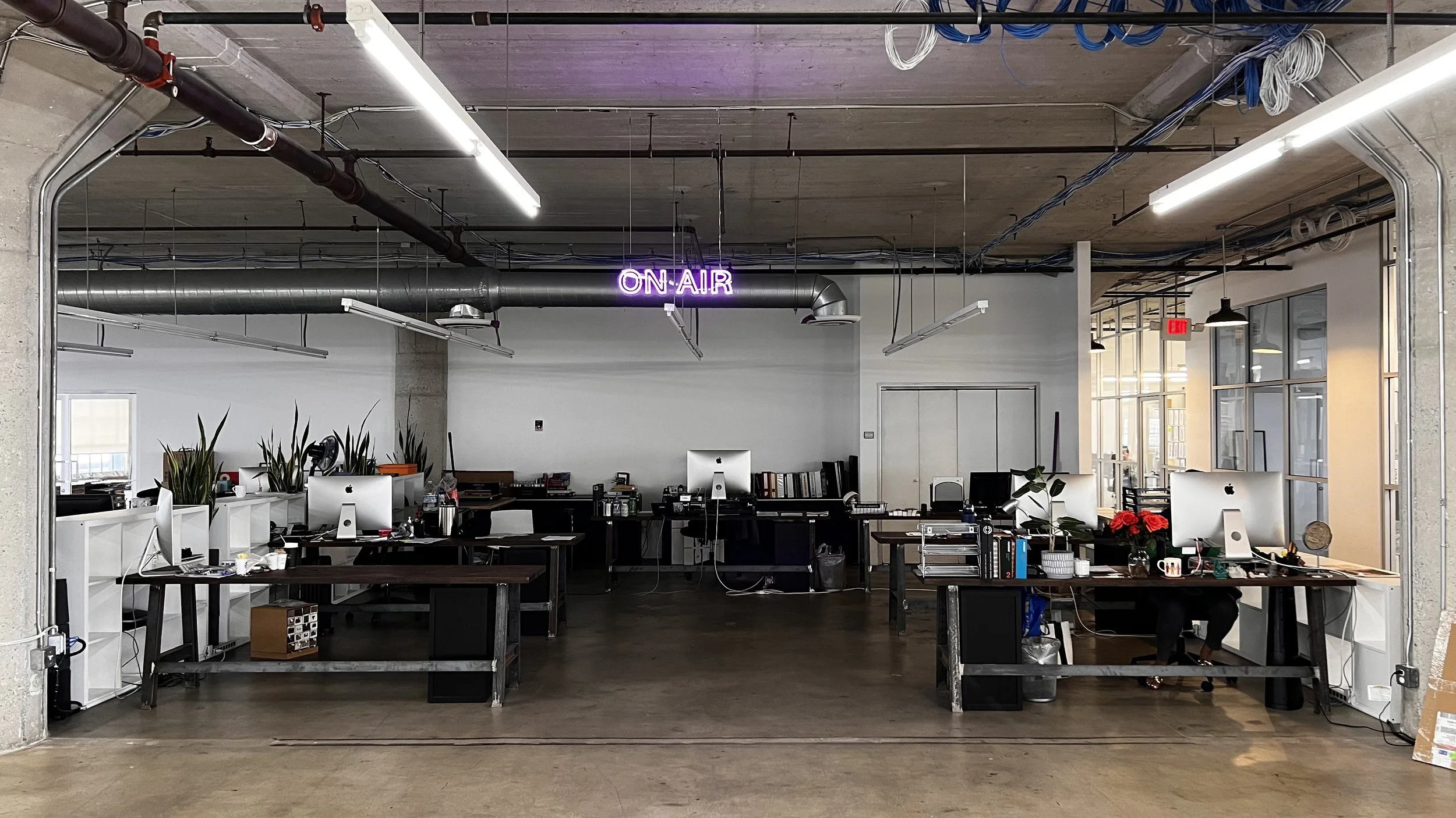 An open office space with multiple desks, computers, and office supplies. There are large windows on the right allowing natural light, and an illuminated neon sign hanging from the ceiling that says "ON-AIR."