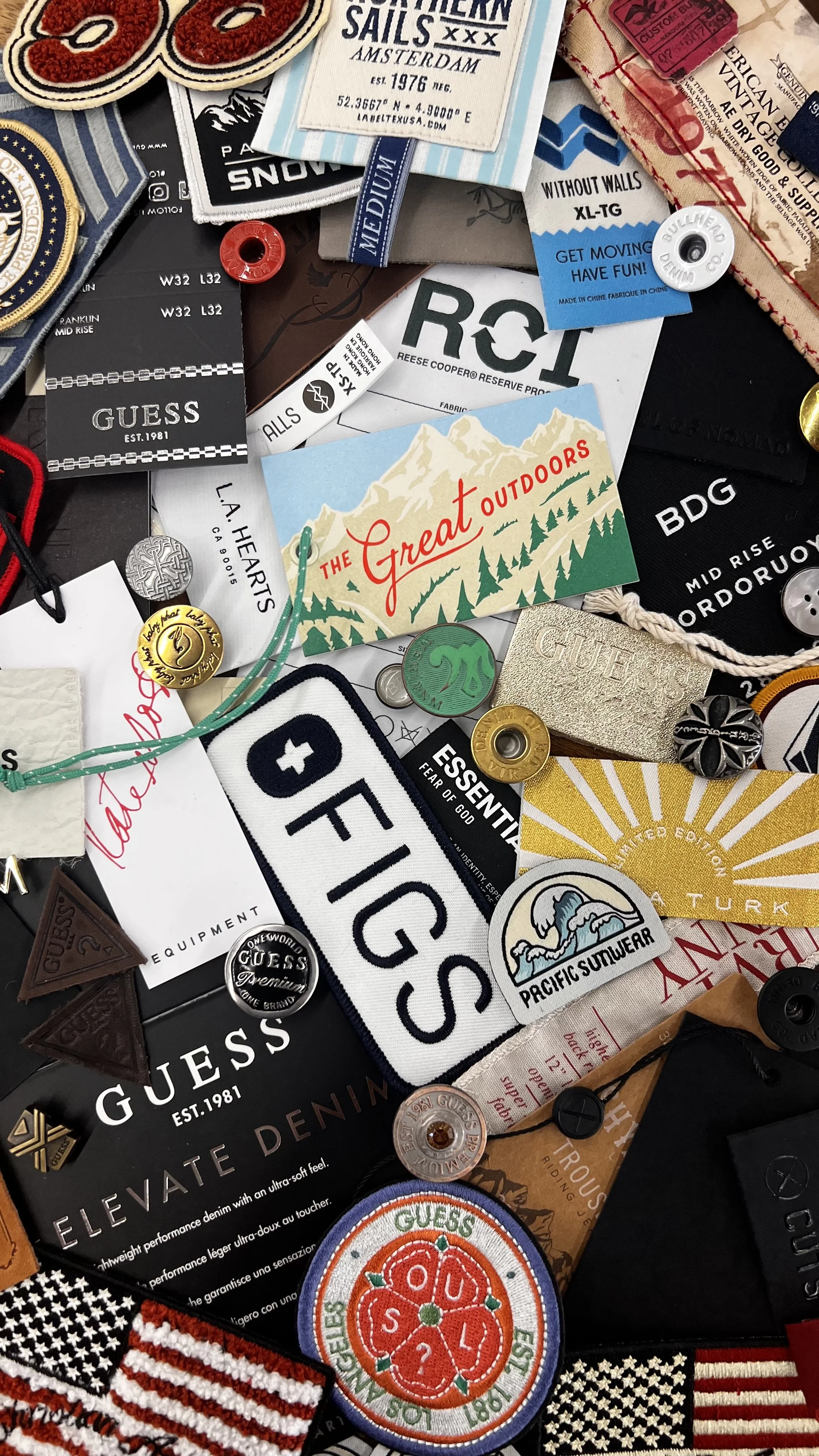 Collage of various patches, buttons, and tags from the Guess brand, including a Pacific Summerwear patch, an American flag patch, and travel-related patches and tags featuring mountains, socks, and outdoor themes.