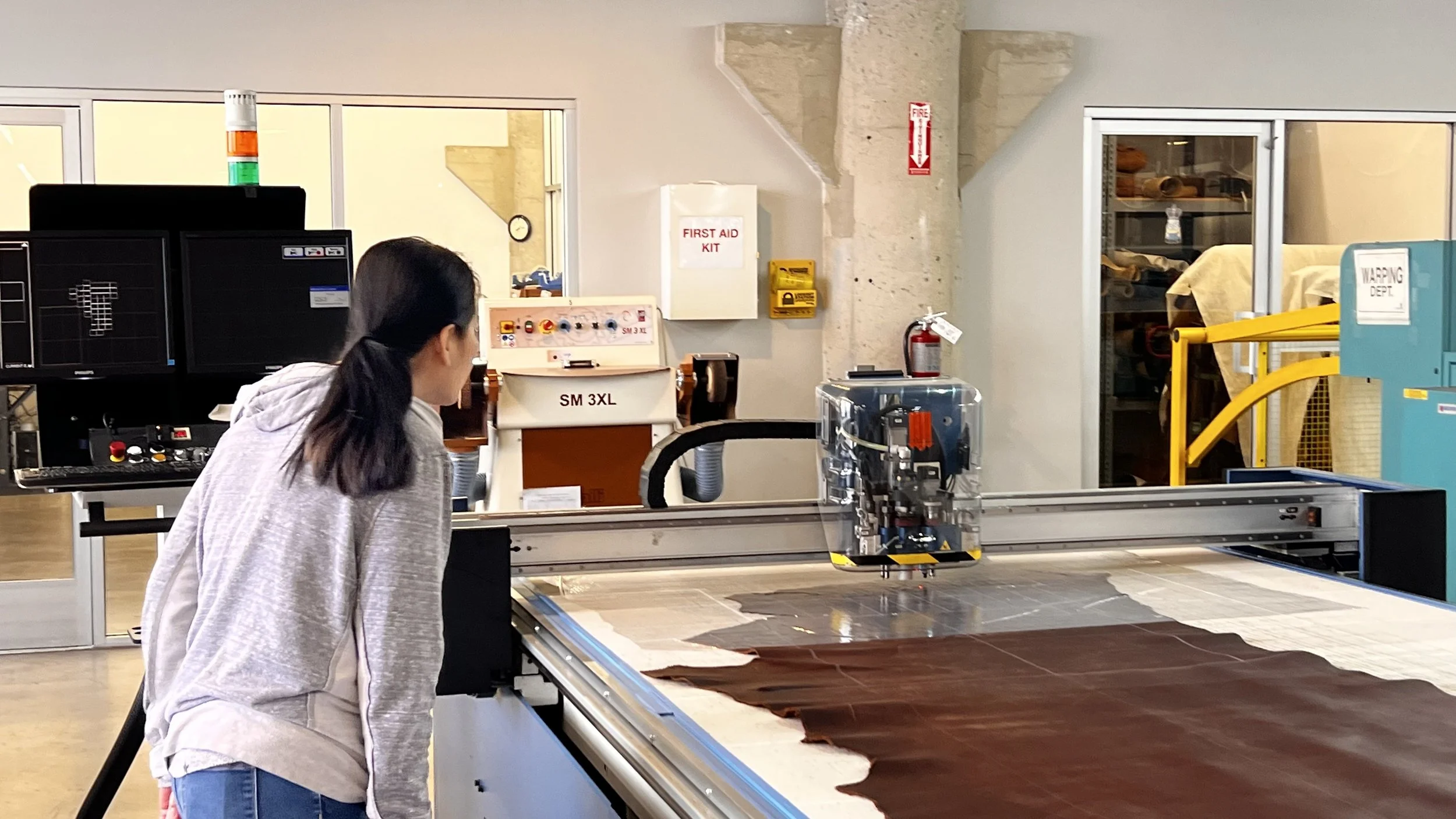 A person operating a CNC machine in a workshop, cutting brown fabric or material, with various tools and safety equipment visible in the background.