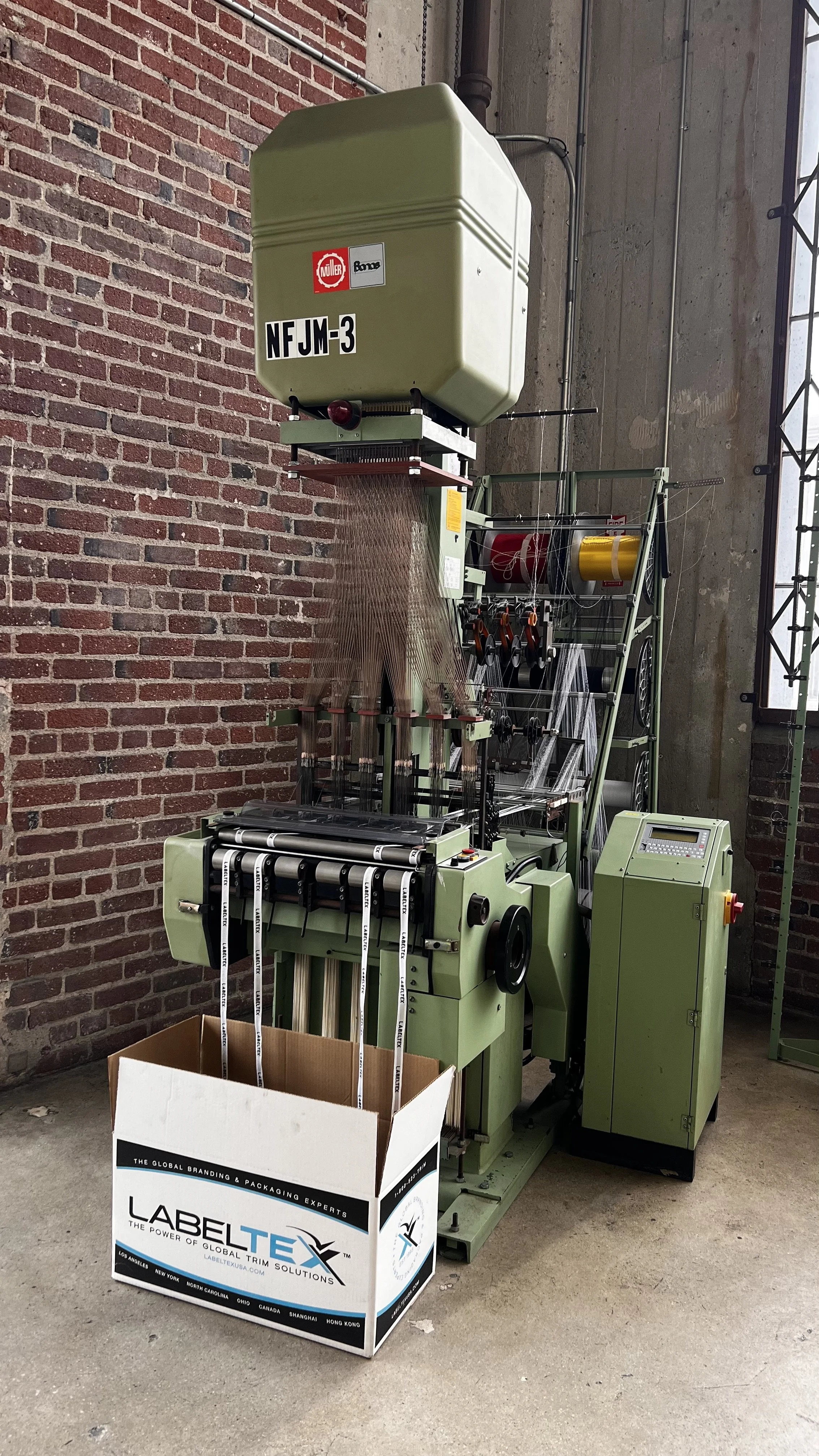 A large green industrial labeling machine printing labels that fall into a box labeled 'LABEL TEX' placed on the floor.