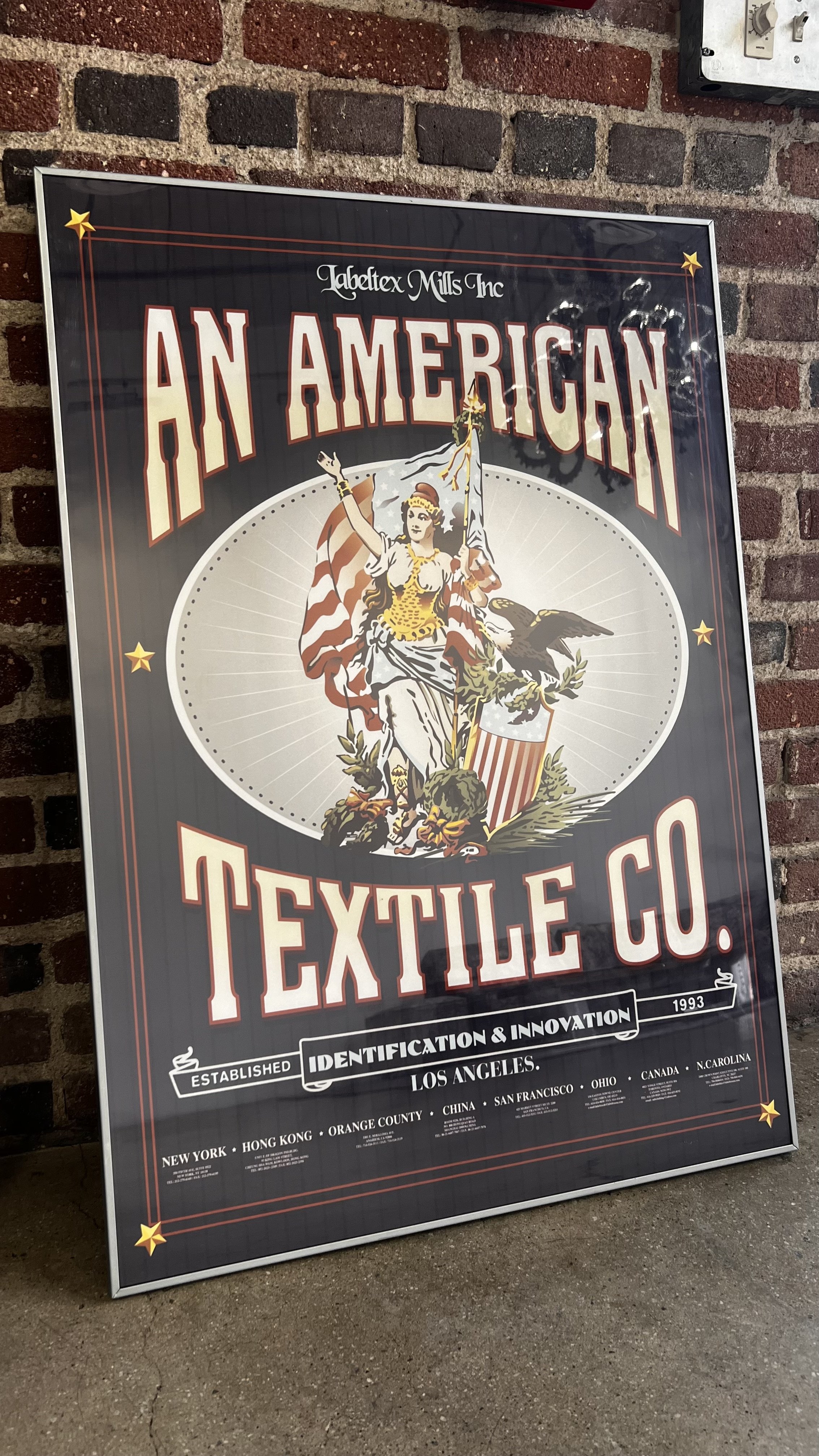 Vintage-style poster for American Textile Co. with an illustration of Lady Liberty holding a flag, an eagle, and shield, set against a radial background, on a dark backdrop with red, white, and beige lettering.