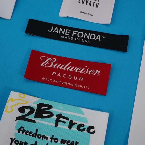 Close-up of four clothing tags on a bright blue background. The tags include brands Jane Fonda, Budweiser Pacsun, and two other labels with partially visible text.