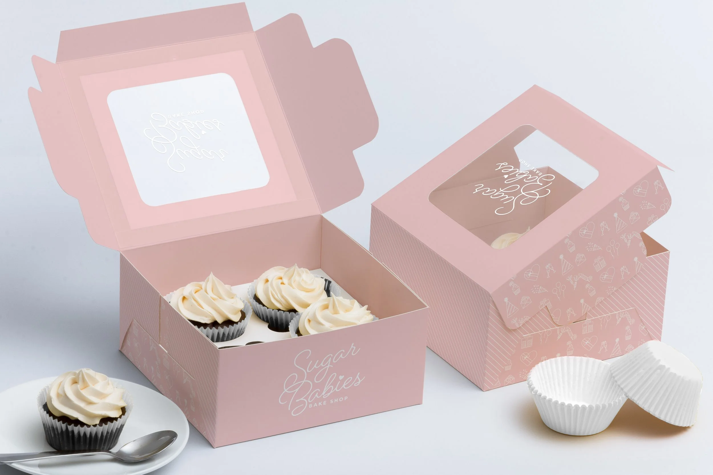 Pink bakery boxes with cupcakes featuring swirled white frosting, some cupcakes are outside the boxes on a white plate, and cupcake liners are stacked nearby.