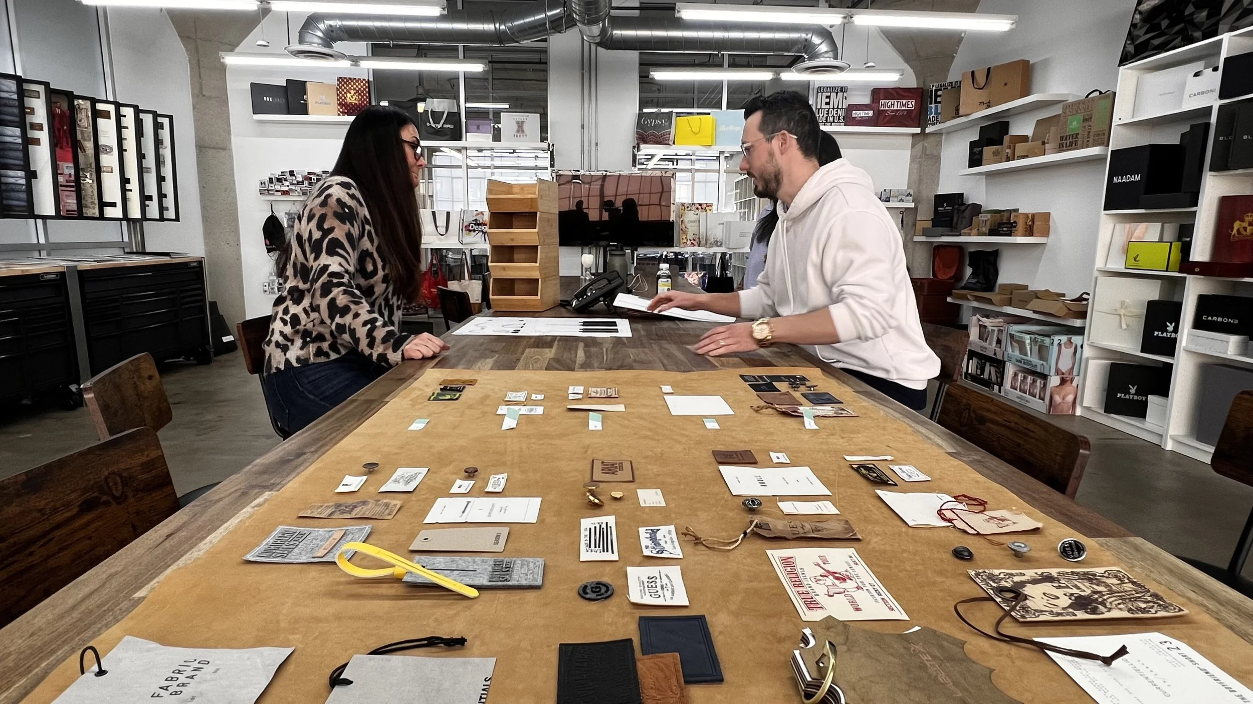 Two people sitting at a wooden table inside a store playing a board game with various game pieces and cards spread out on the table.