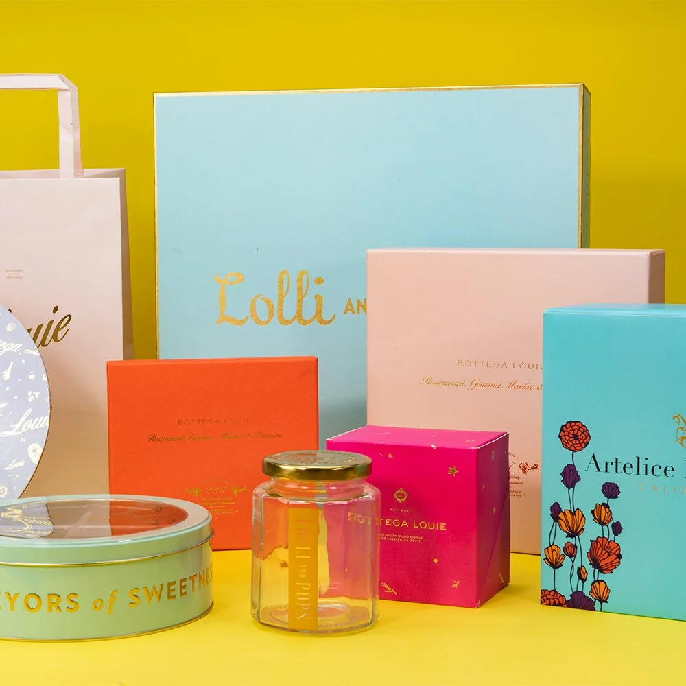 Assorted colorful designer packaging and containers on a yellow surface against a yellow wall, including boxes and a glass jar.