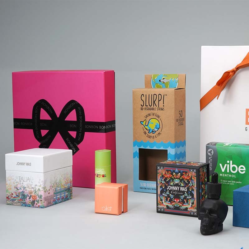 Arrangement of various packaged products including gift boxes, a box of biodegradable straws labeled SLURP, a box of Vibe Menthol vape pods, and decorative items on a plain background.