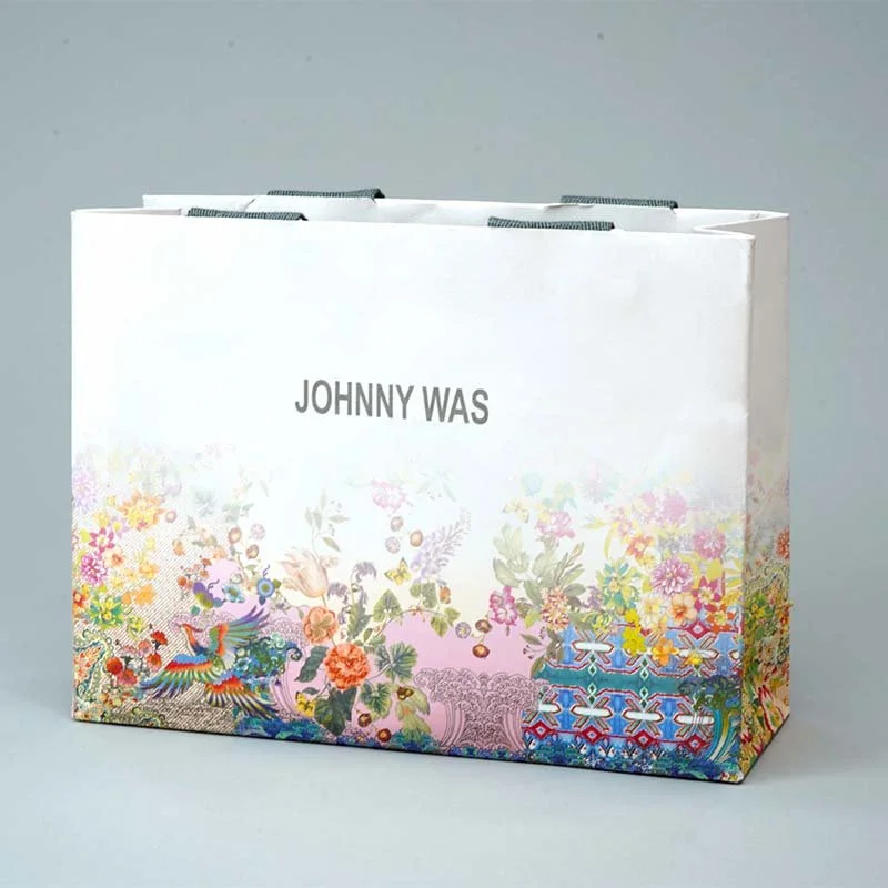 A white gift bag with a floral pattern at the bottom and the words "JOHNNY WAS" printed on it, set against a plain gray background.