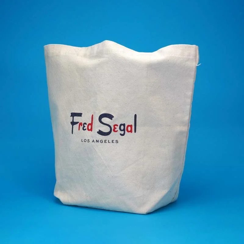 A beige tote bag with 'Fred Segal Los Angeles' printed on it, standing against a blue background.