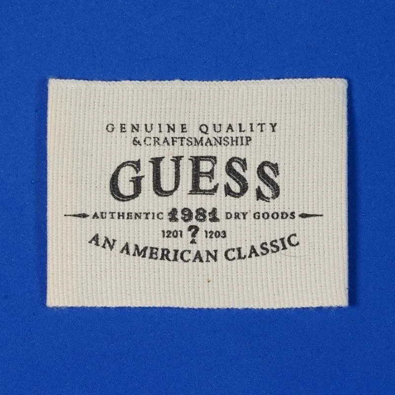 Close-up of a white fabric label with black text reading "Genuine Quality & Craftsmanship Guess Authentic 1981 Dry Goods An American Classic" against a blue background.